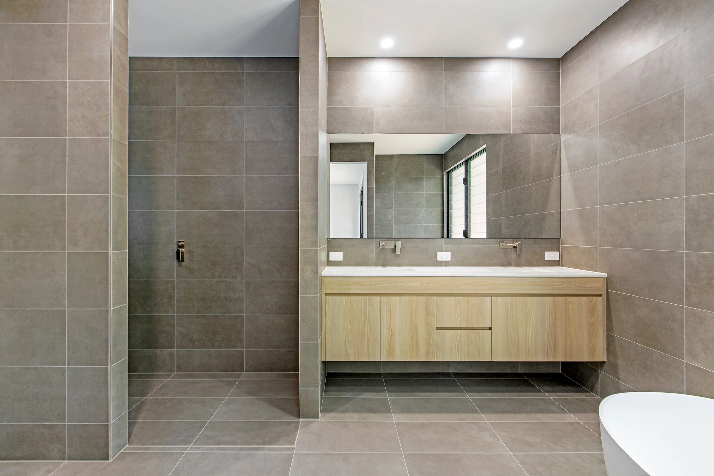 Modern bathroom with gray tiles on walls and floor, a large mirror above a light wooden vanity, a window with blinds, and recessed ceiling lights.