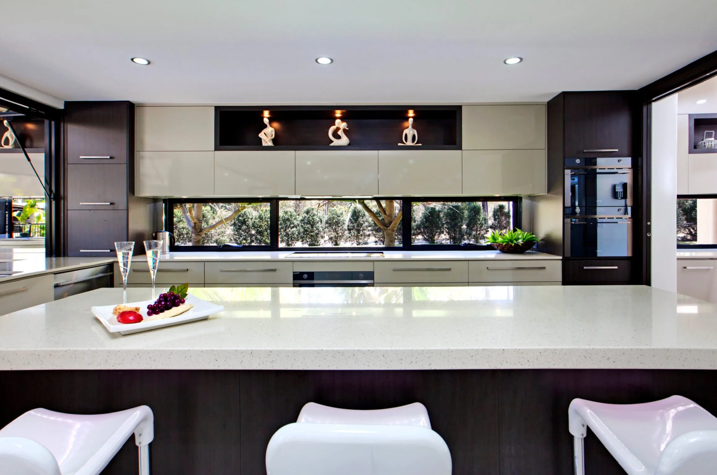 Modern kitchen with dark wood and beige cabinets, large windows showing trees outside, white countertop with a fruit dessert, and white chairs around a dining table.