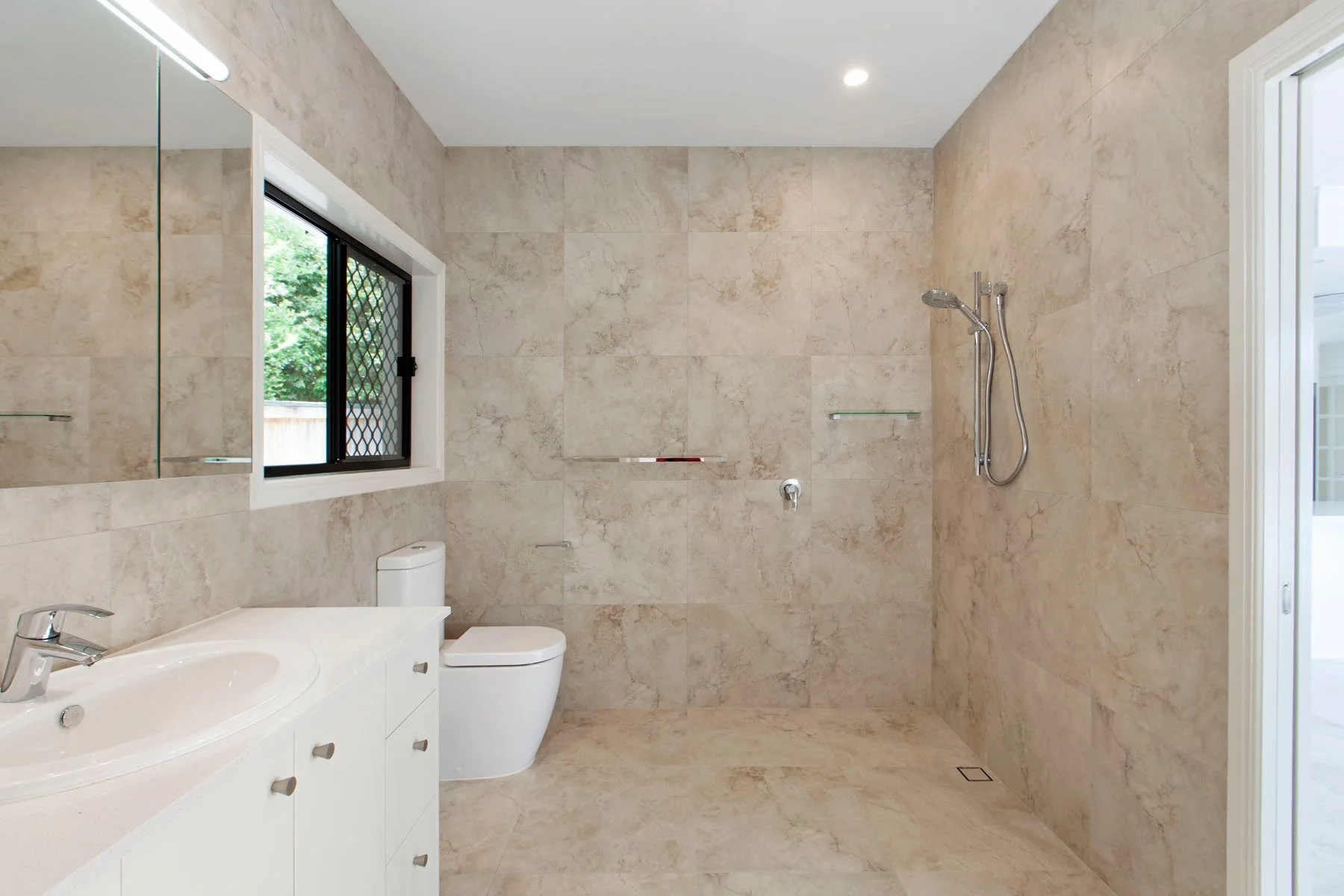 Bathroom with beige marble walls and floor, a white sink vanity, a toilet, and a walk-in shower with a showerhead and glass shelf. There is a window with a black security grille, and a mirror above the sink.