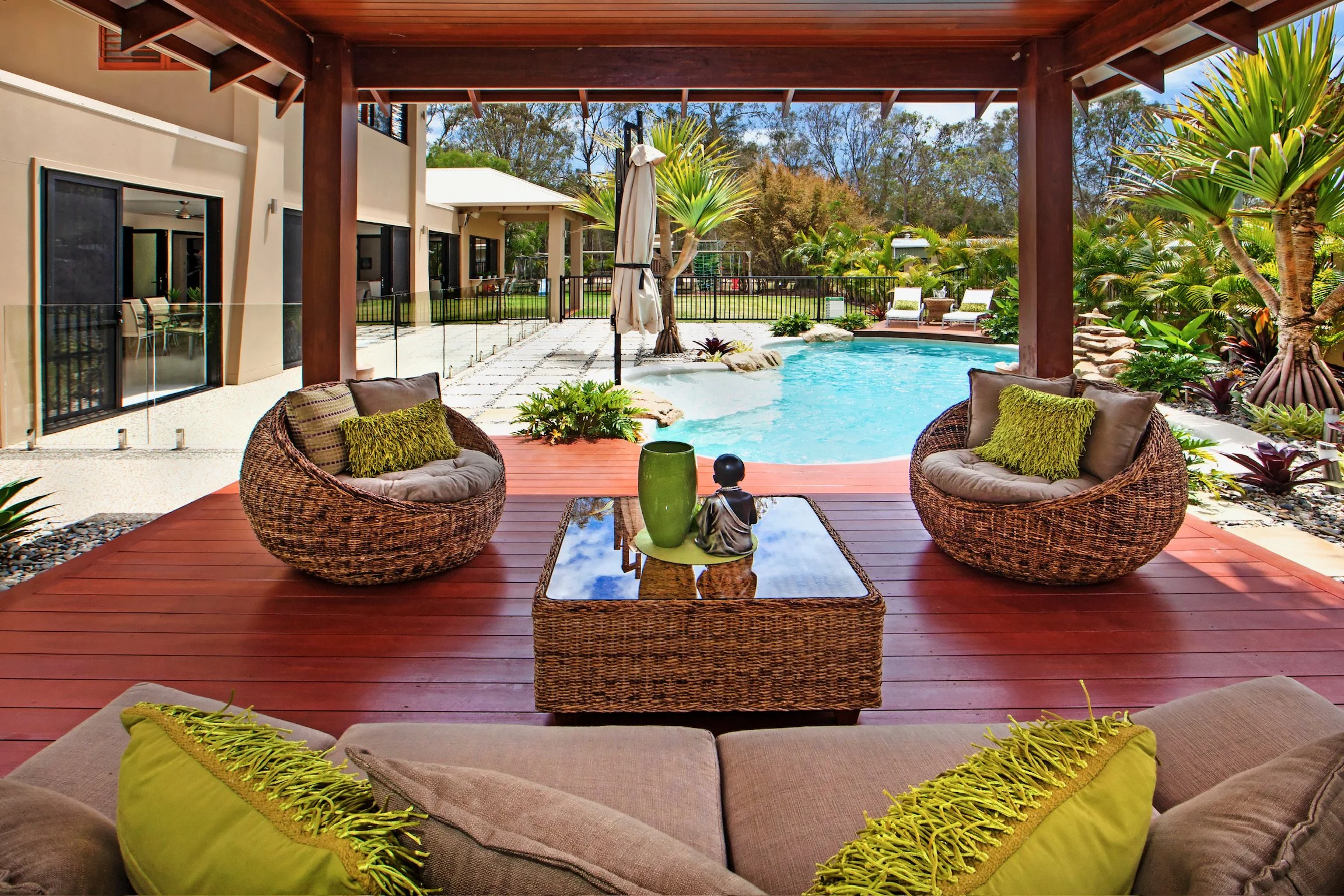 View of a backyard pool area, seen from a covered patio with rattan chairs and a glass-topped table, surrounded by lush green tropical plants and trees.