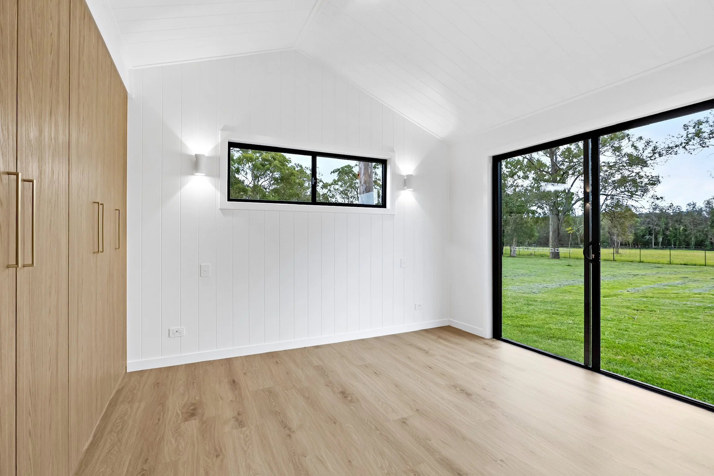 Empty room with white walls, wooden floors, a large sliding glass door opening to a green yard, and a small window with trees outside.