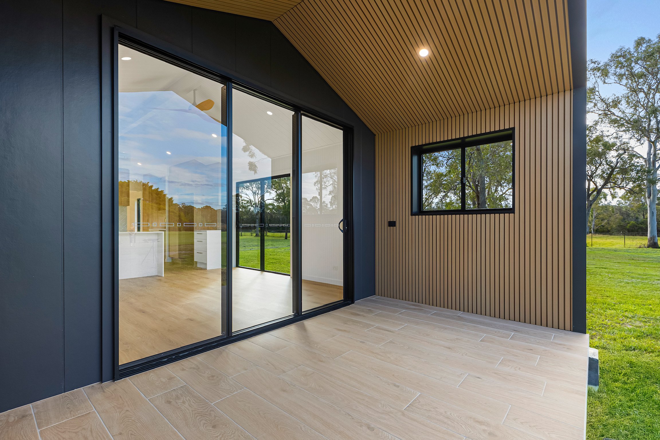 A modern house with large glass sliding doors, wooden paneling on the exterior, and a view of green trees and grass outside.