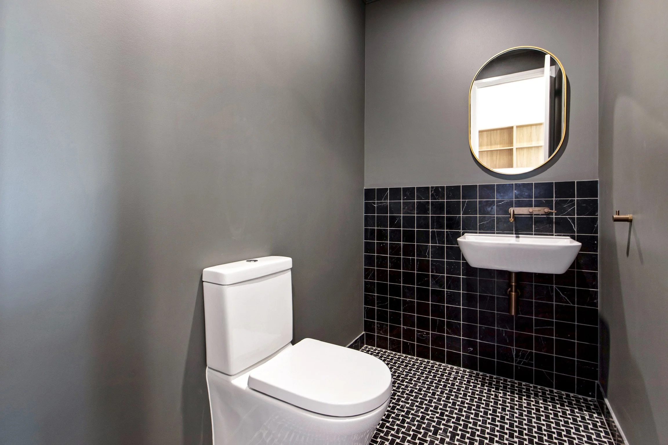 Modern bathroom with a gray wall, black tiled accent wall, white toilet, small white sink with gold fixtures, a round mirror with gold trim, and a window with wooden shelves visible through the mirror.