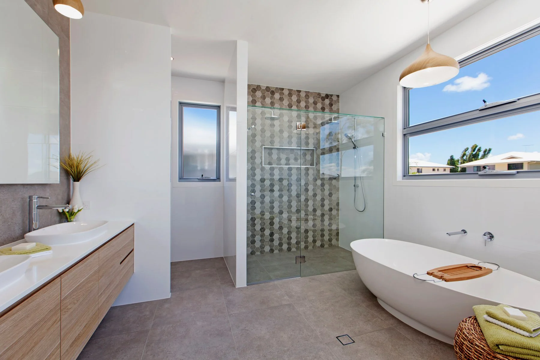 Modern bathroom with a large window showing blue sky and trees, featuring a walk-in glass shower with hexagonal tile wall, a freestanding bathtub with a wooden bath tray, a wooden vanity with two vessel sinks, and a wicker basket with towels.
