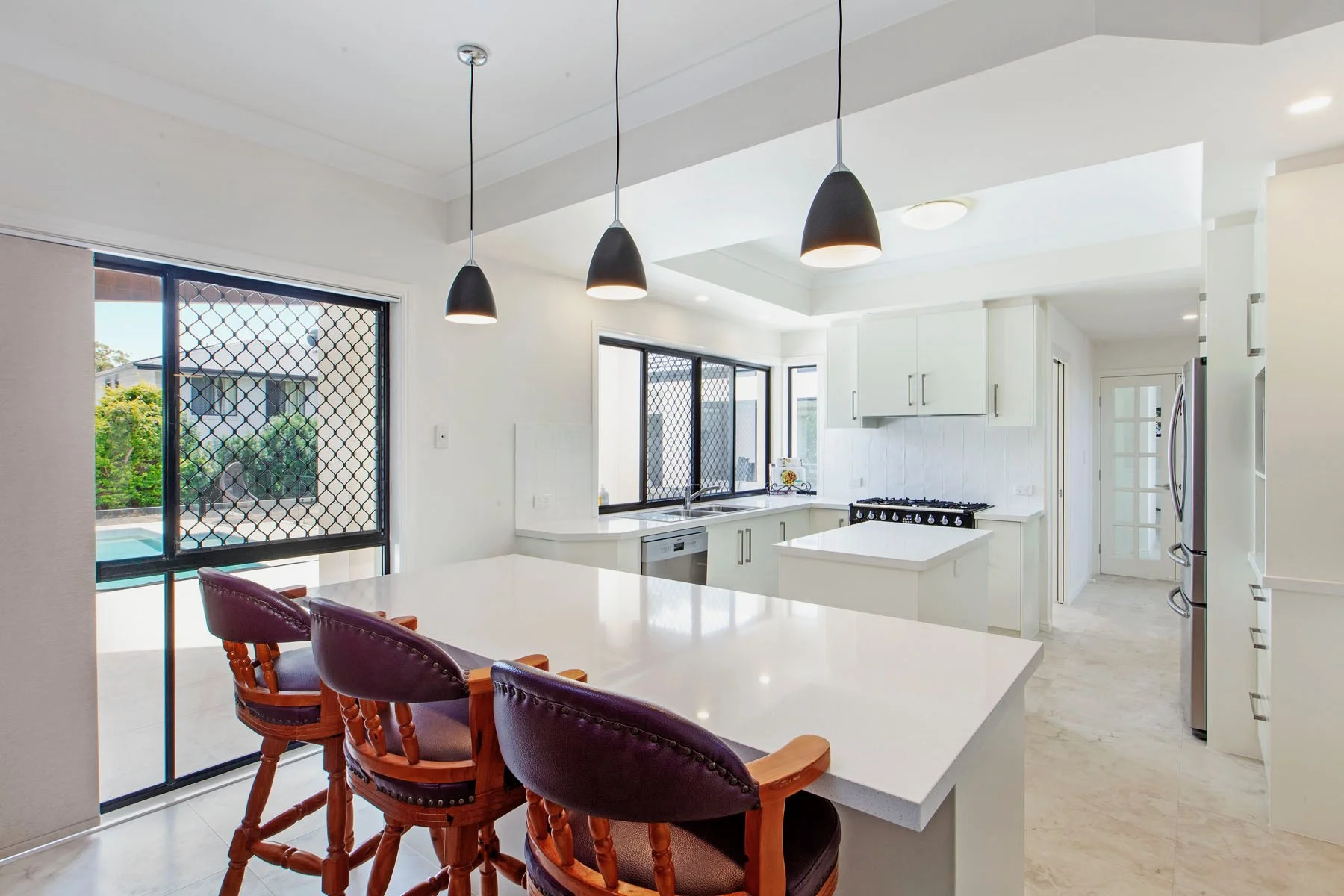 Modern kitchen with white cabinetry, black pendant lights, stainless steel appliances, large windows with security screens, and a breakfast bar with purple cushioned chairs.