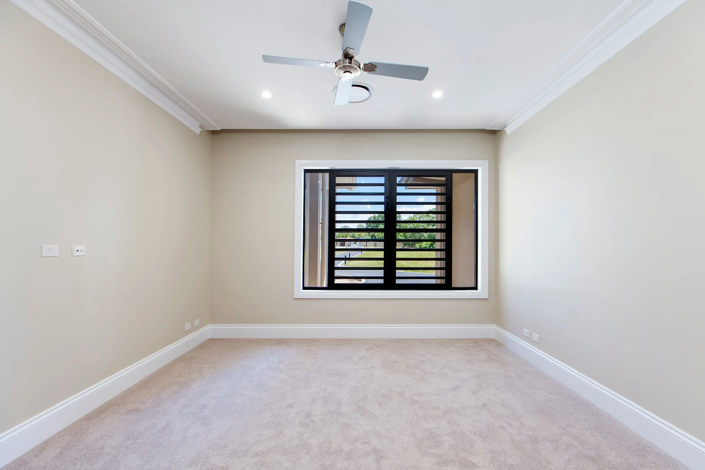 Empty room with beige walls, white crown molding, beige carpeting, a window with black horizontal blinds, and a ceiling fan with four blades.
