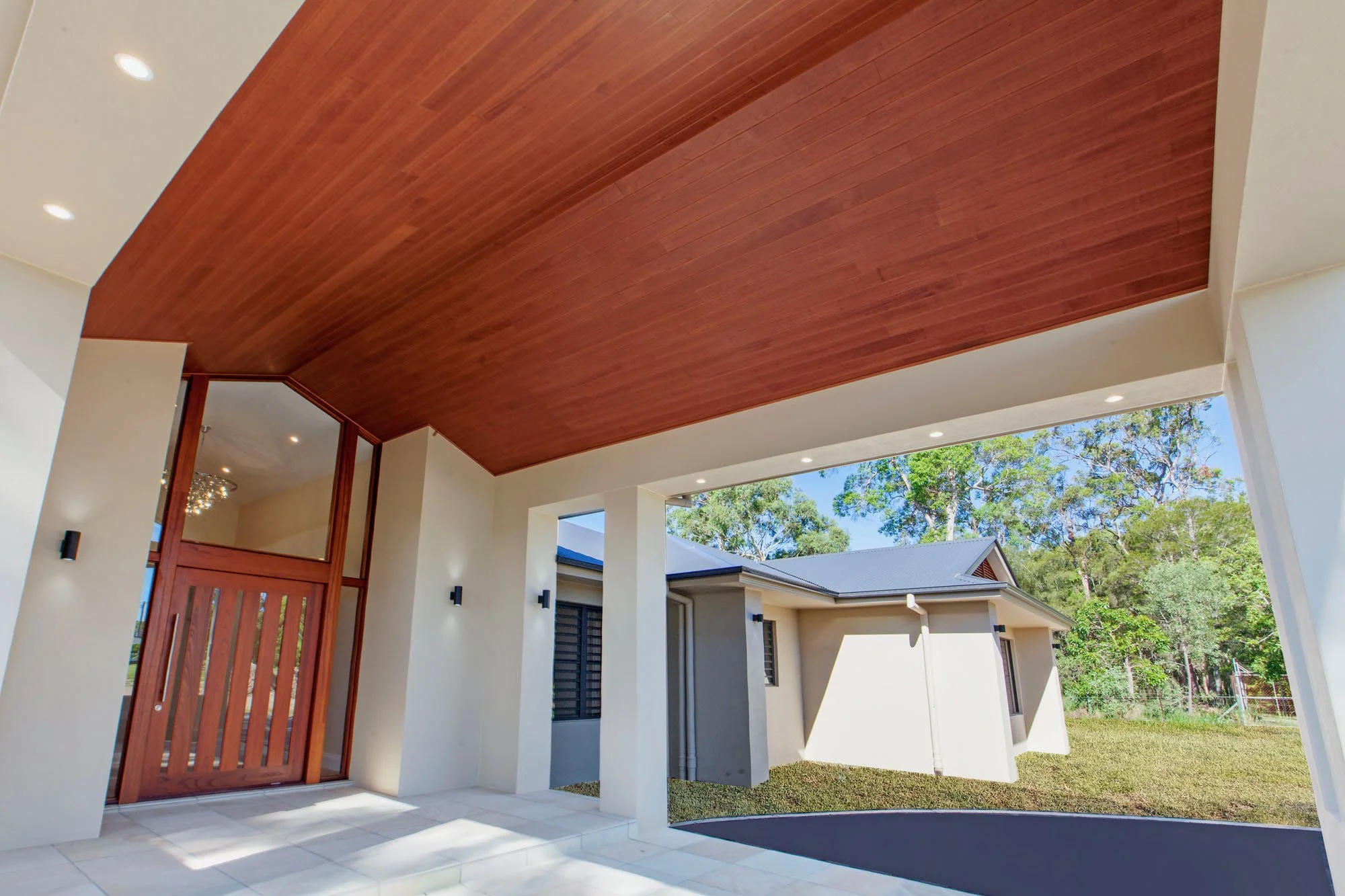 Modern house exterior with white walls, a wooden ceiling over the porch, and black exterior light fixtures, set against a background of trees and a clear sky.