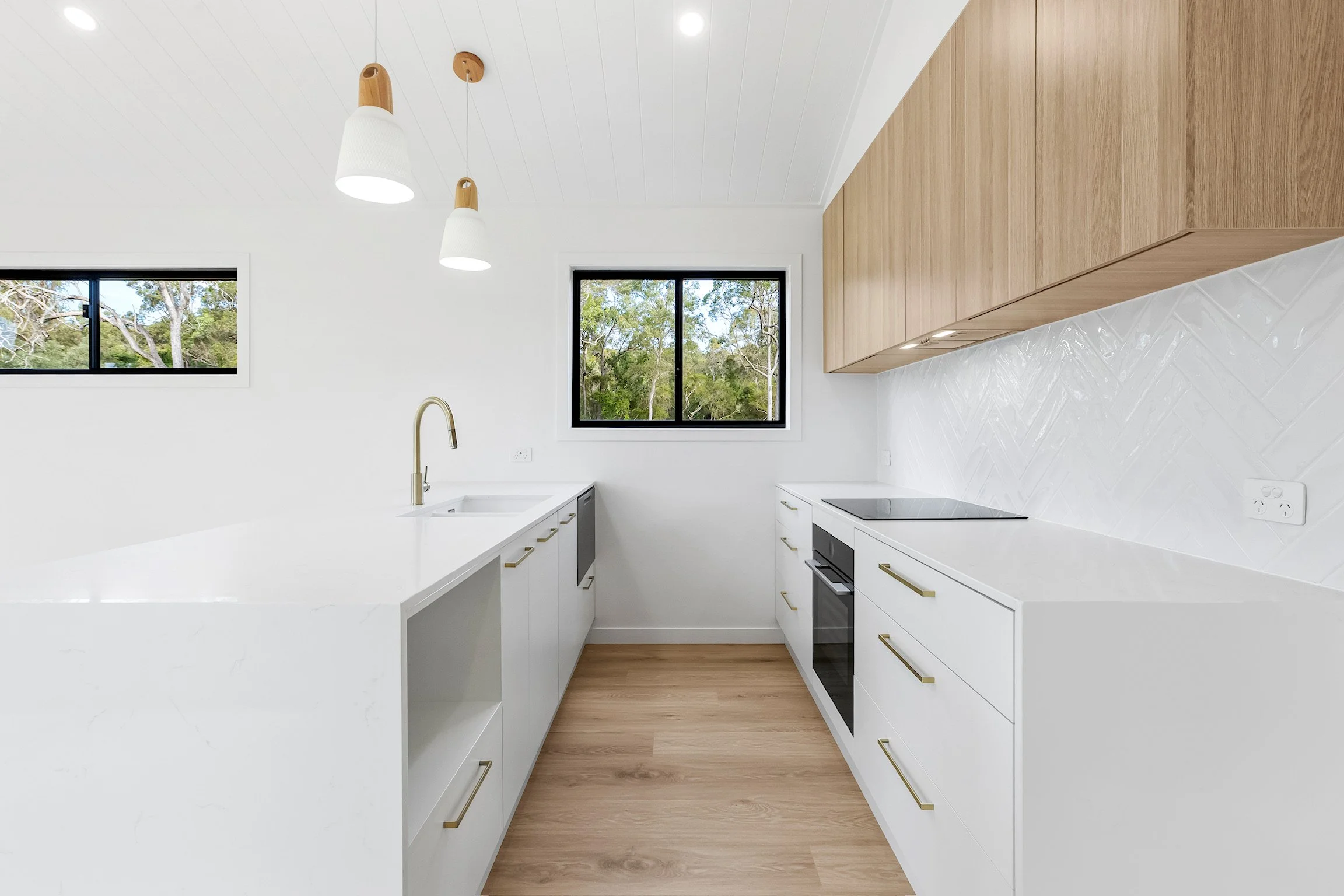 Modern kitchen with white cabinets, wooden upper cabinets, a white countertop, a gas stove, a stainless steel sink, two windows showing trees outside, and ceiling pendant lights.