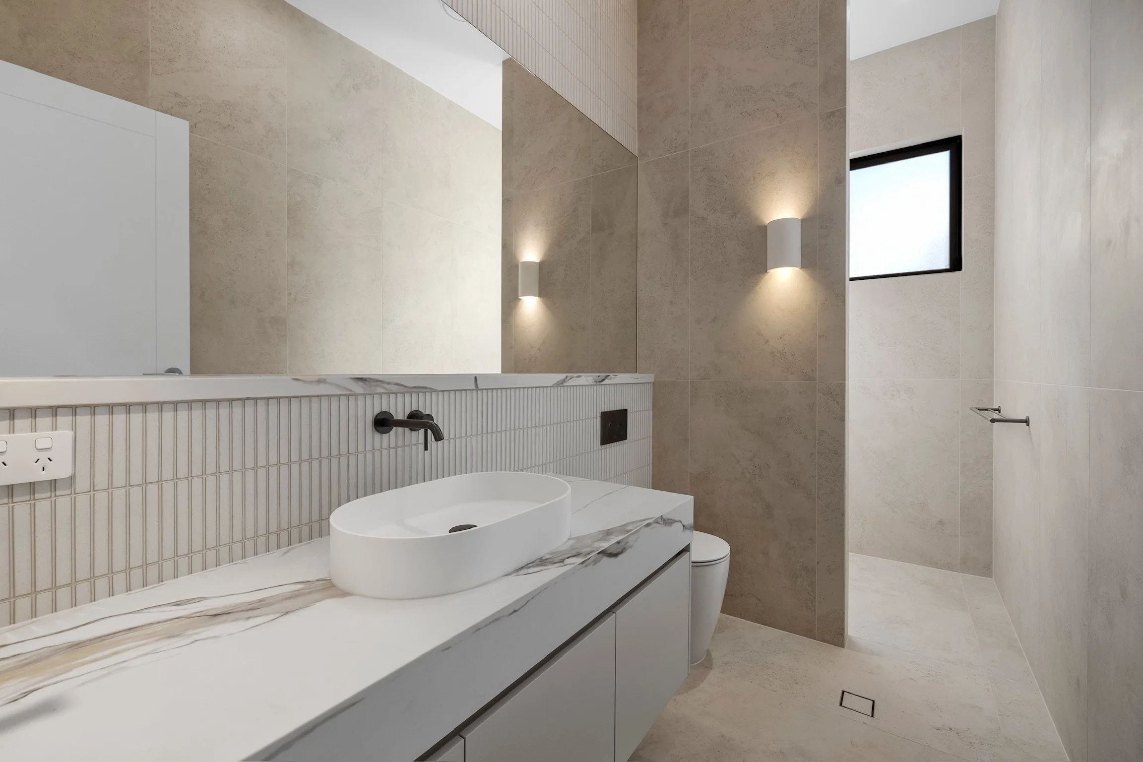 A modern bathroom with beige tiled walls, a white vanity with a marble countertop, a vessel sink, a wall-mounted faucet, a large mirror, wall sconces, a small window, and a towel holder.