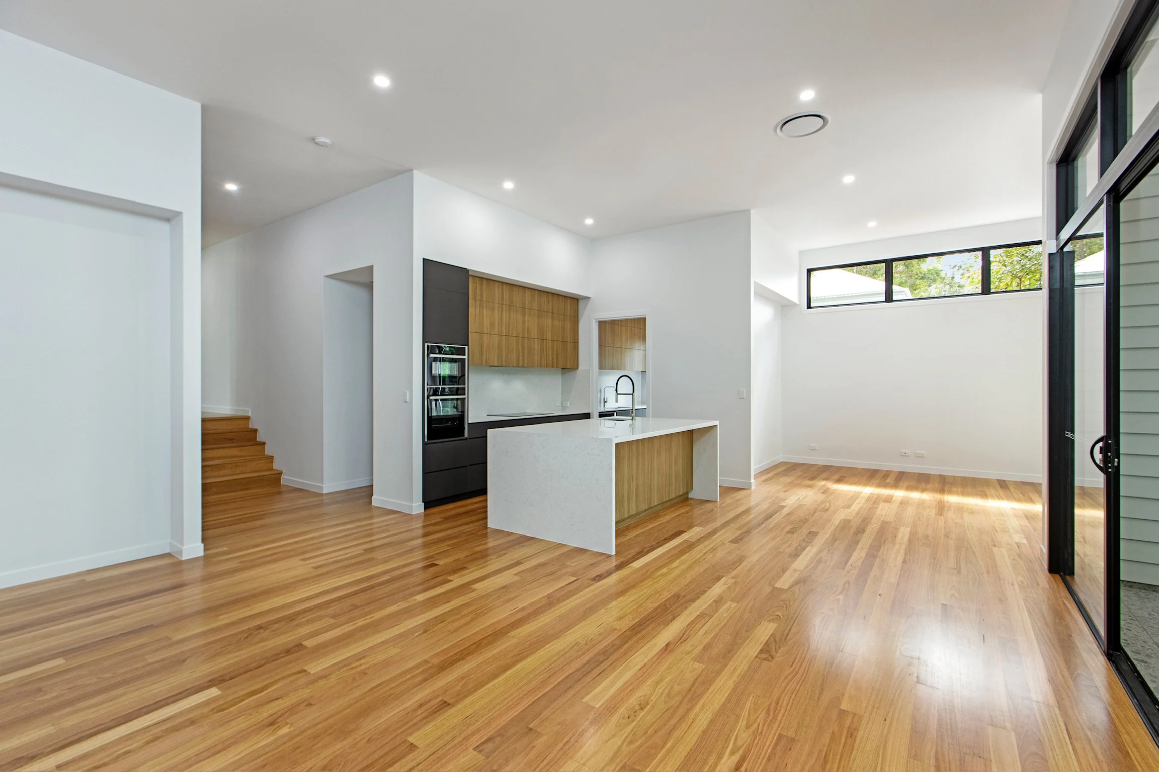 Interior view of a modern, spacious room with hardwood floors, white walls, a kitchen with wooden cabinets, a white island, and large windows letting in natural light.