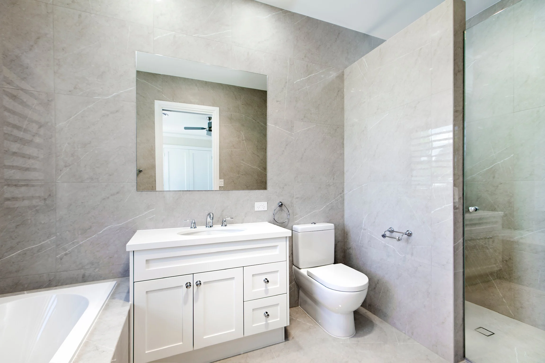Modern bathroom with beige marble walls and floor, white vanity with two sinks, a large mirror above, toilet, and shower area with glass door.