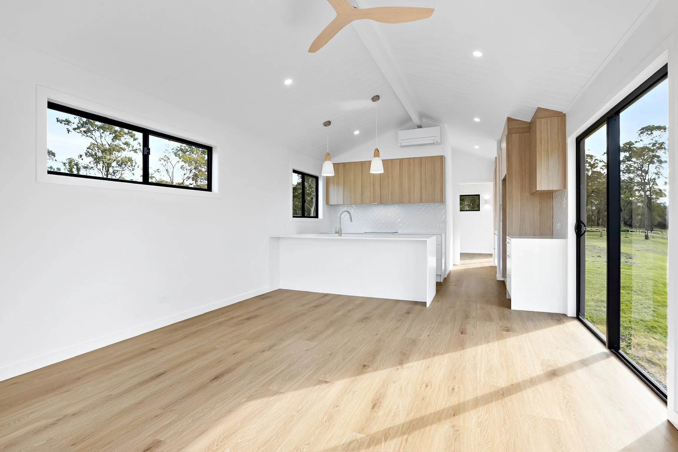 Interior of a modern, minimalistic kitchen with light wood flooring, white walls, large windows, and black-framed sliding glass doors that overlook a green outdoor landscape. The ceiling has recessed lighting and a ceiling fan.