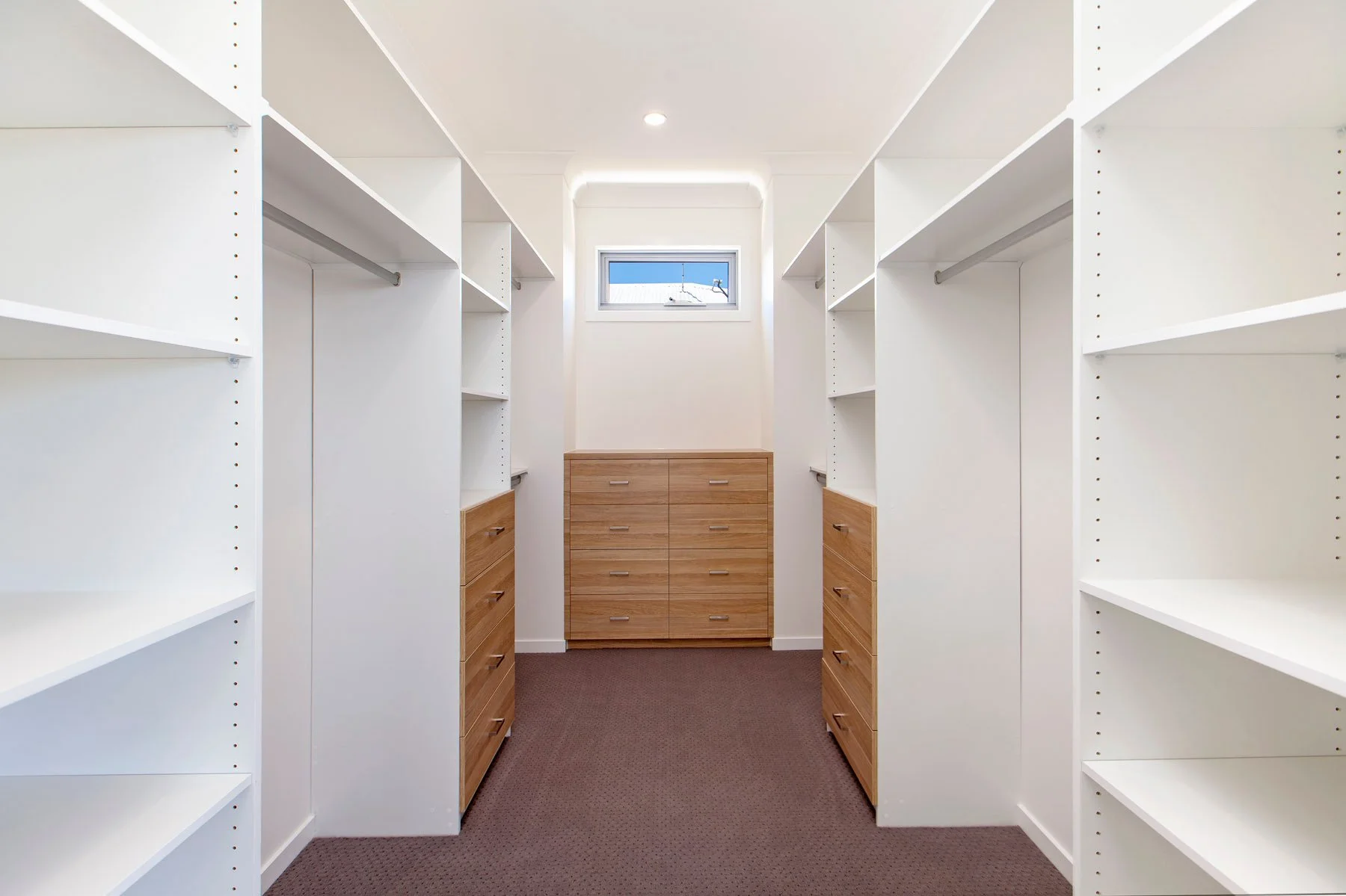 Empty walk-in closet with white shelves and drawers made of light wood, small window at the back letting in natural light, dark carpet flooring.