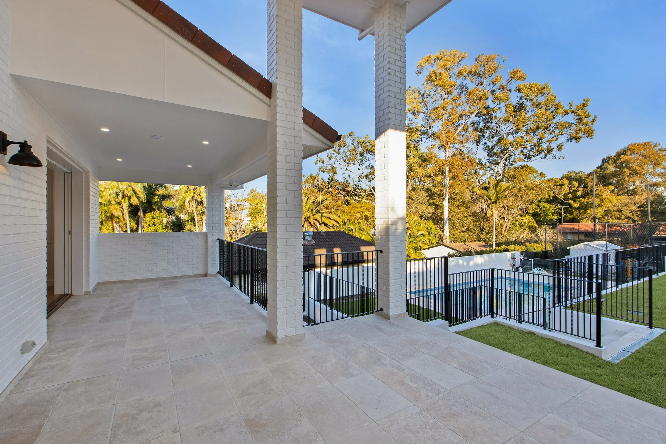 Balcony with tiled floor, white brick pillars, black metal railing, and a view of a backyard with trees, a swimming pool, and neighboring houses under a blue sky.