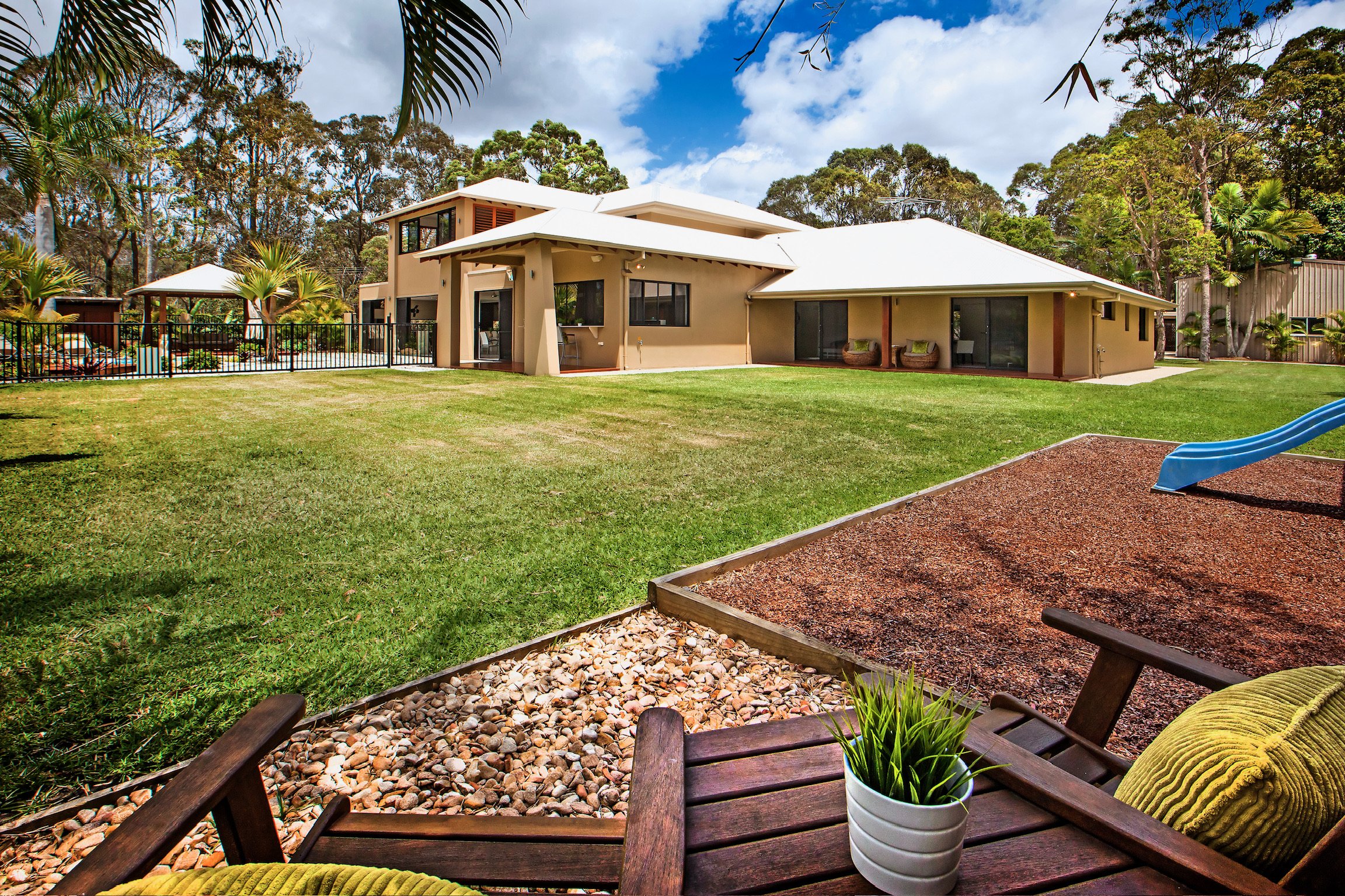 A modern house with large windows, beige walls, and a white roof, set in a lush green yard with a lawn, trees, and a small playground featuring a blue slide. The foreground shows a wooden table with a yellow cushion and a potted plant, while the back