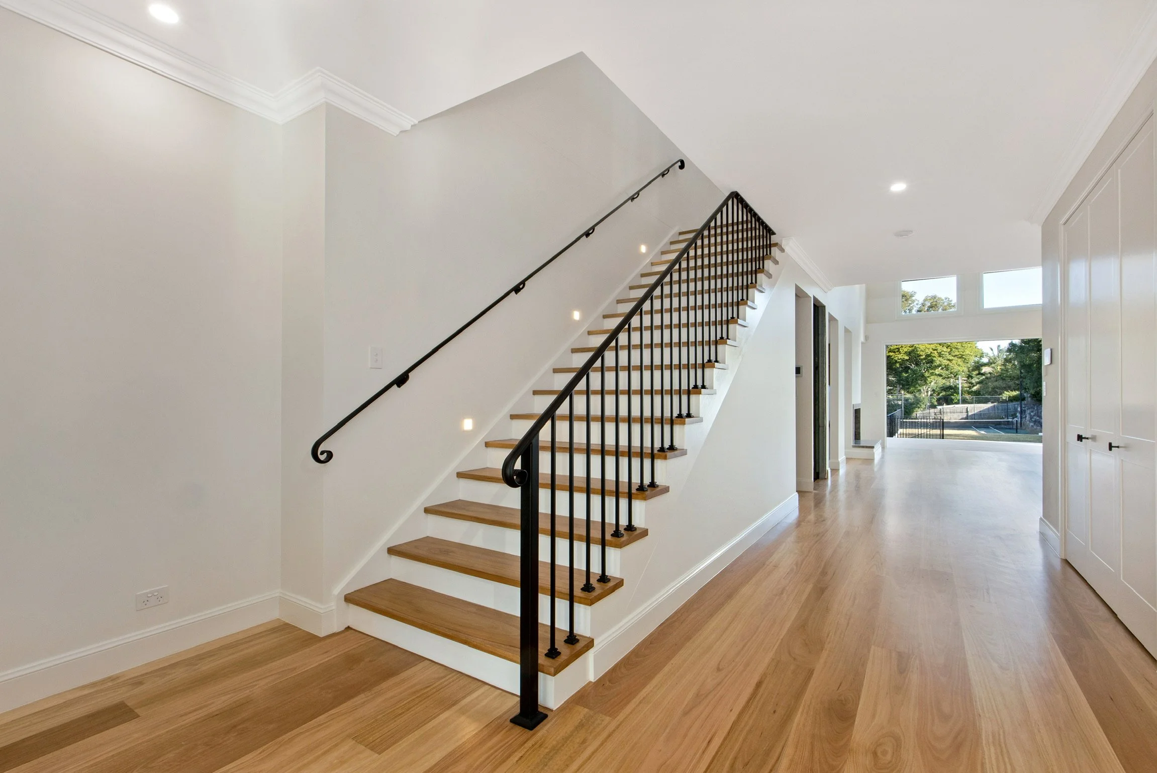 Interior view of a modern home with a staircase featuring wooden steps and black metal railing, natural light coming through windows, light-colored walls, and hardwood floors.