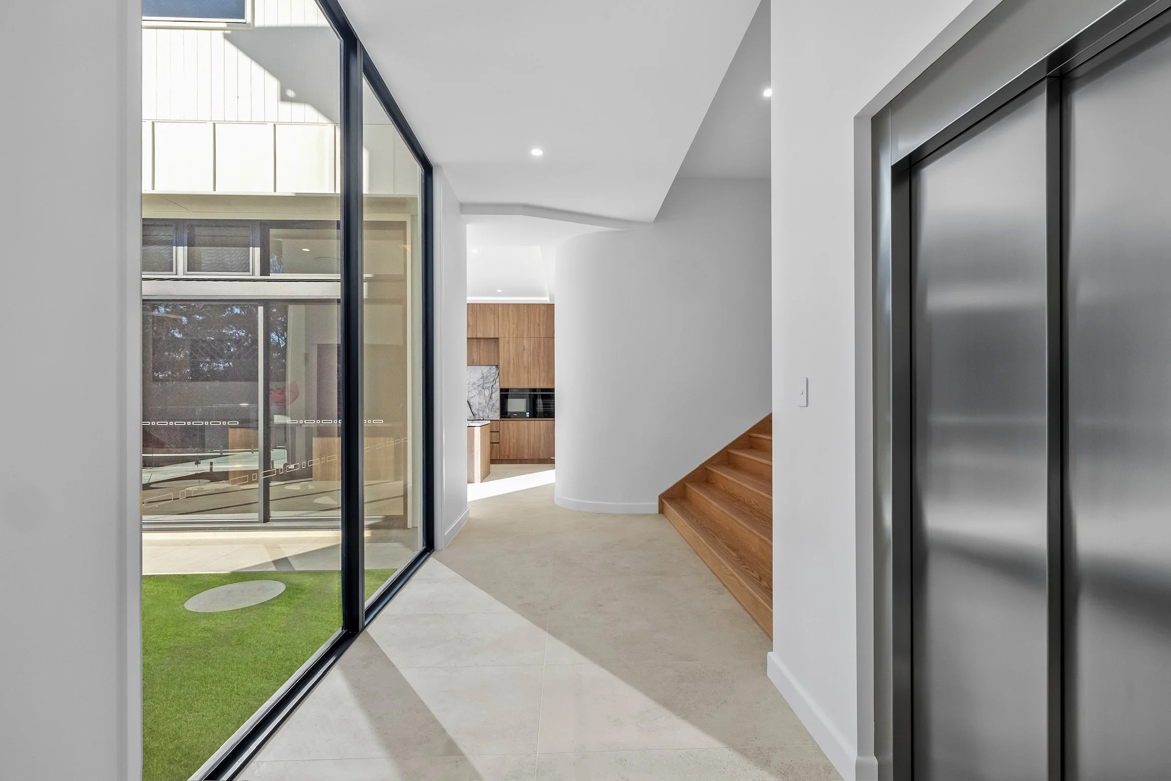 Modern interior space with an open view of a kitchen, wooden stairs, glass door, and elevator door.