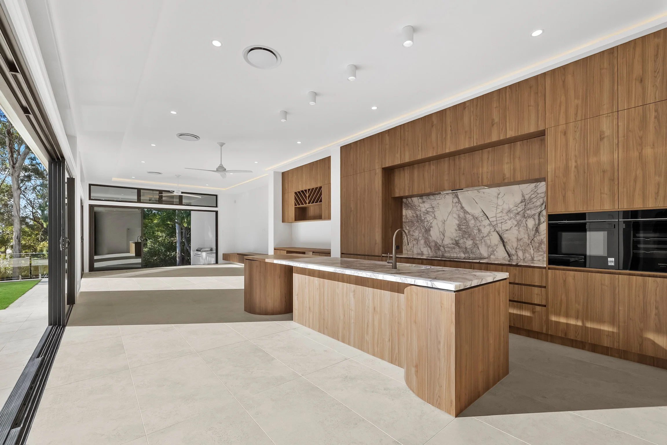 Modern kitchen with wooden cabinets, marble backsplash, island with marble countertop, and large windows overlooking green trees outside.