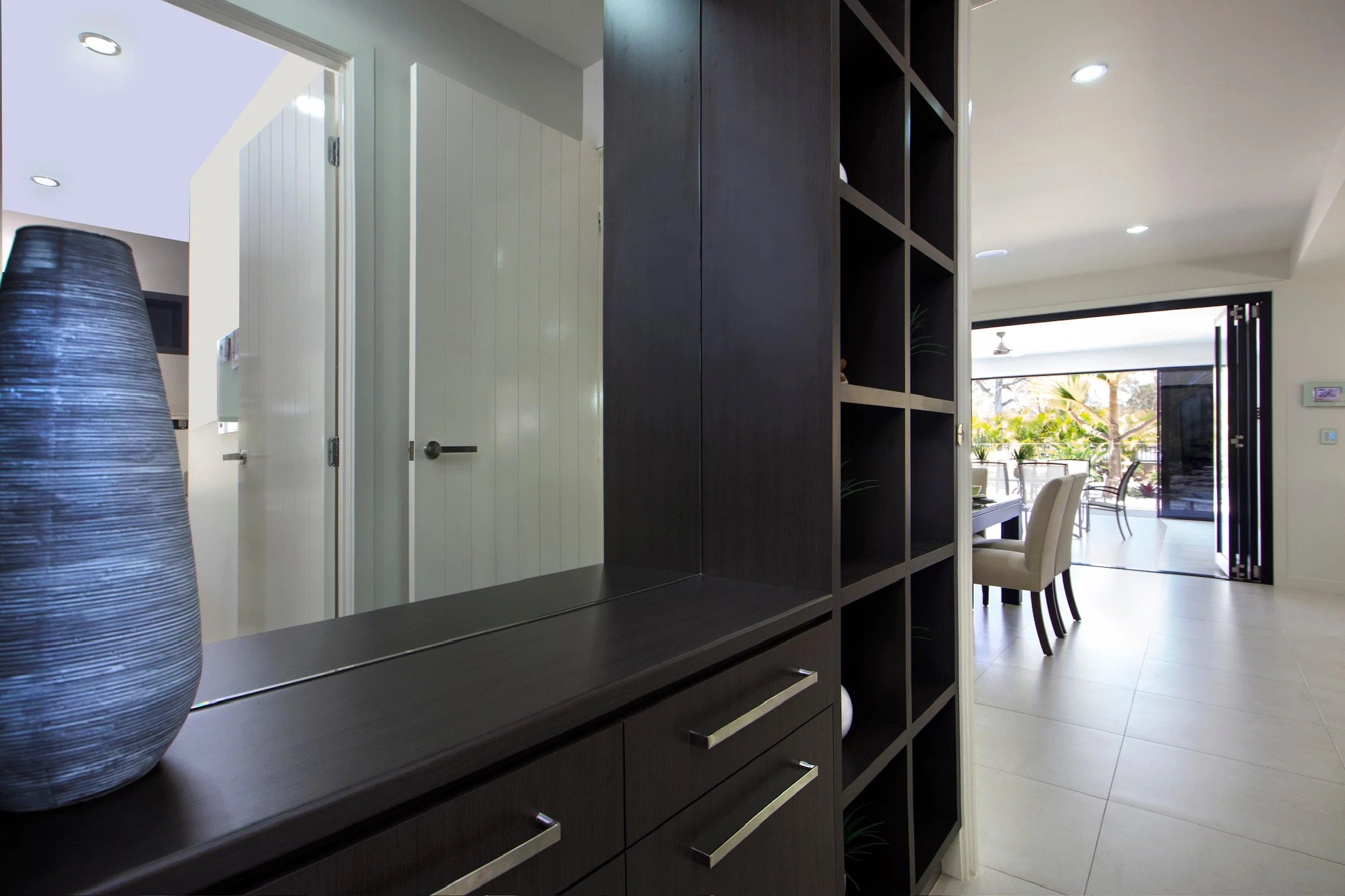 Interior view of a modern living space with a dark wooden shelving unit, a dining area with a table and chairs, and large sliding glass doors opening to a patio with outdoor furniture and greenery.