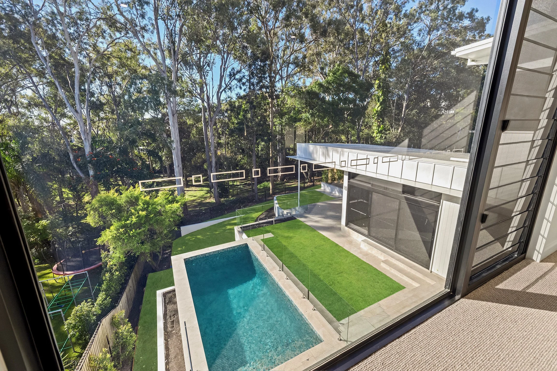 A view from an open window showing a backyard with a swimming pool, a green lawn, trees, and part of a modern house with large glass sliding doors.