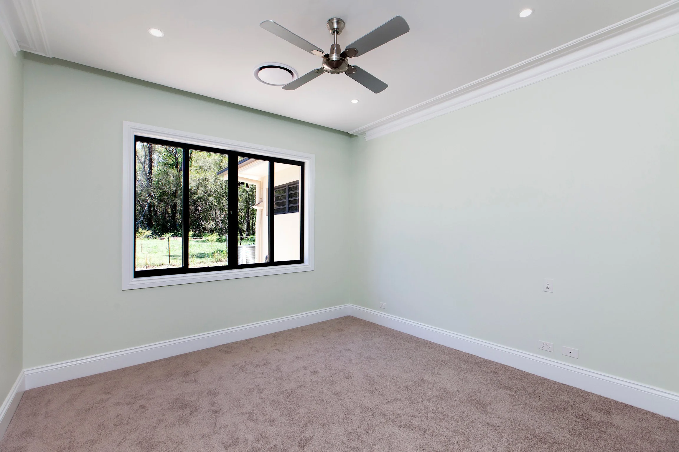 Empty room with beige carpet, light green walls, large window showing green trees outside, ceiling fan, and recessed lighting.