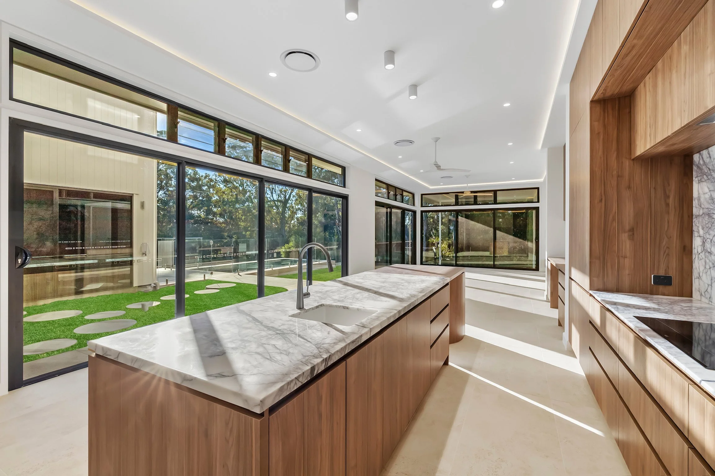 Modern kitchen with marble countertops, wooden cabinets, large glass windows, and a view of the garden outside.
