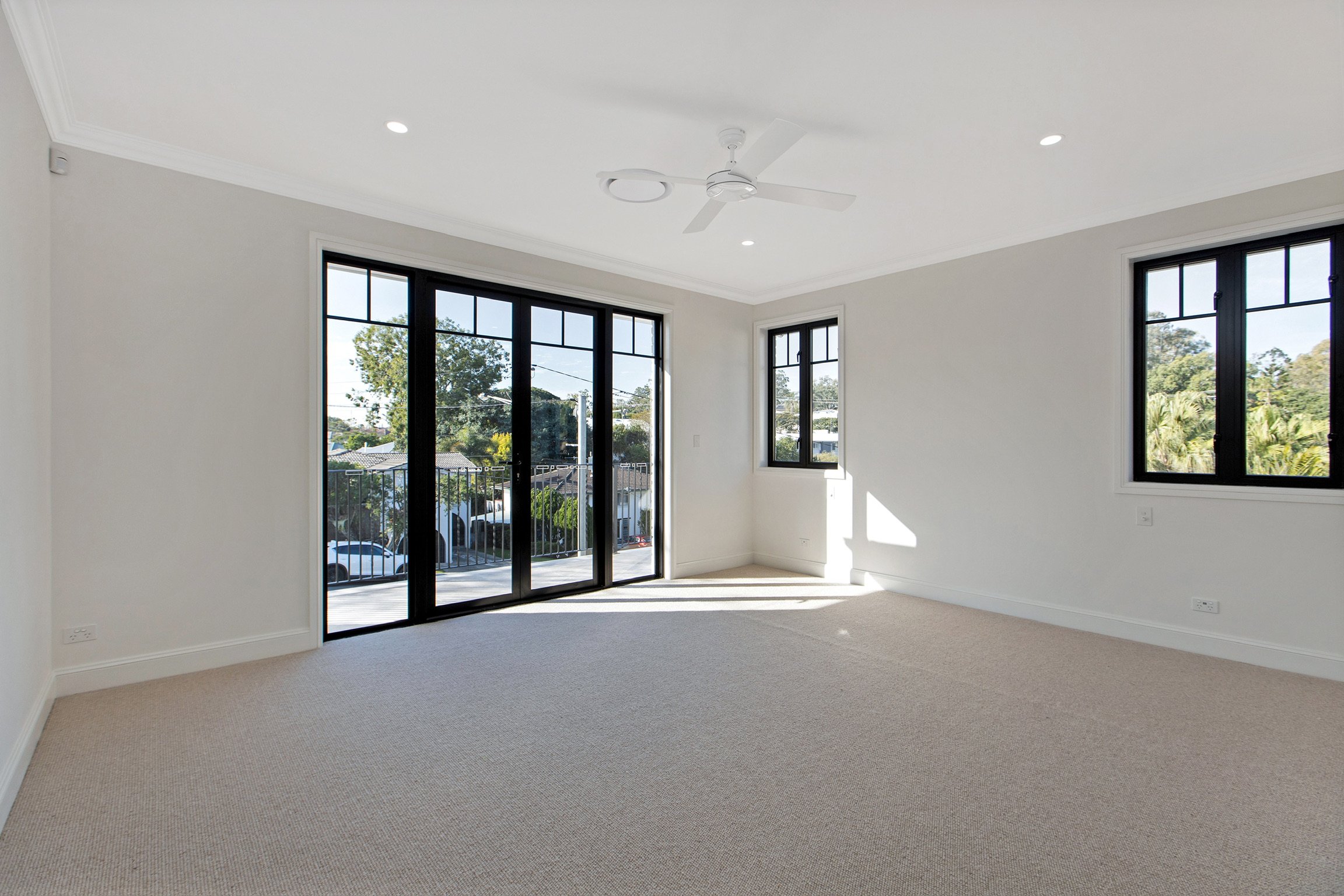 Empty room with beige carpet, white walls, large black-framed windows, and sliding glass door leading outside, with trees and houses visible through the windows.