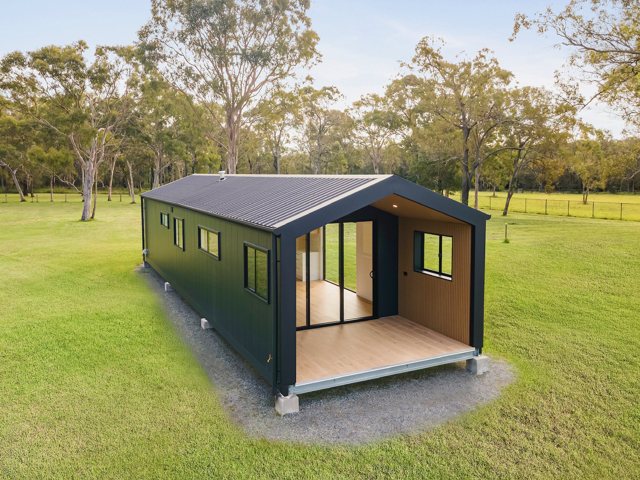 A small modern house with black exterior walls and a sloped metal roof is on a large grassy field with trees in the background. The house has large windows and an open interior space visible from outside.