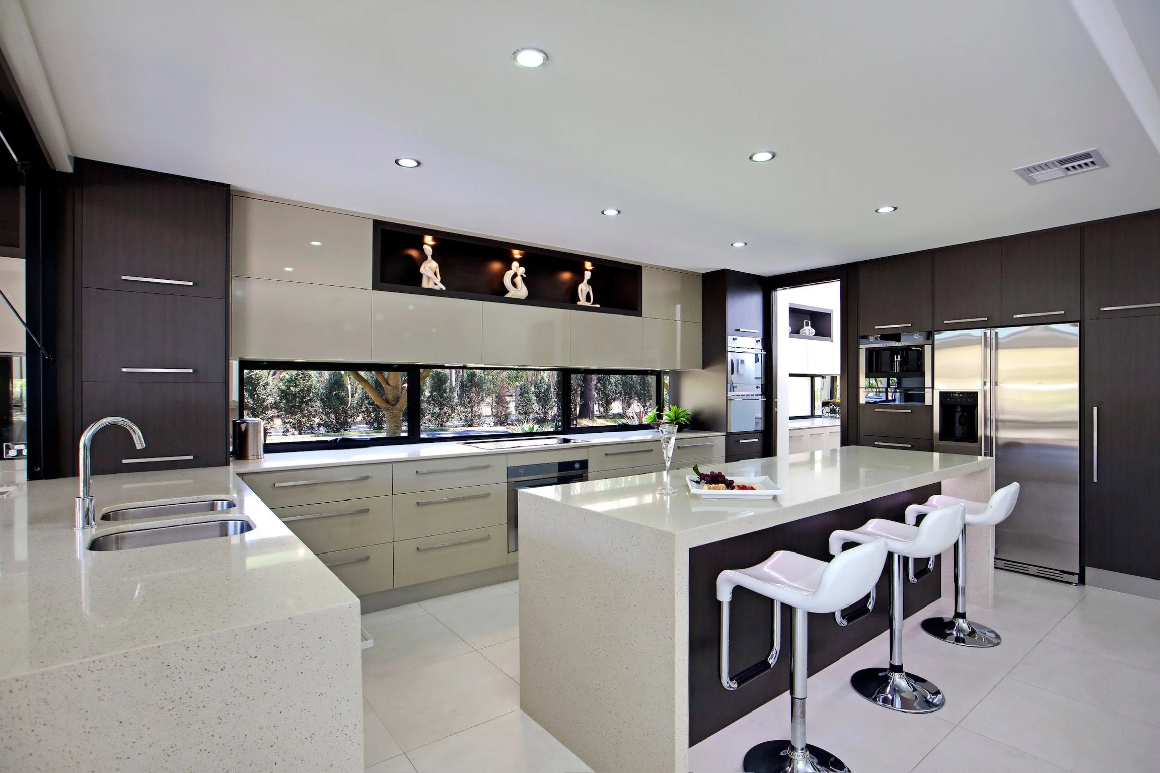 Modern kitchen with white countertops, dark wood cabinets, bar stools, and decorative sculptures on a shelf near a large window.