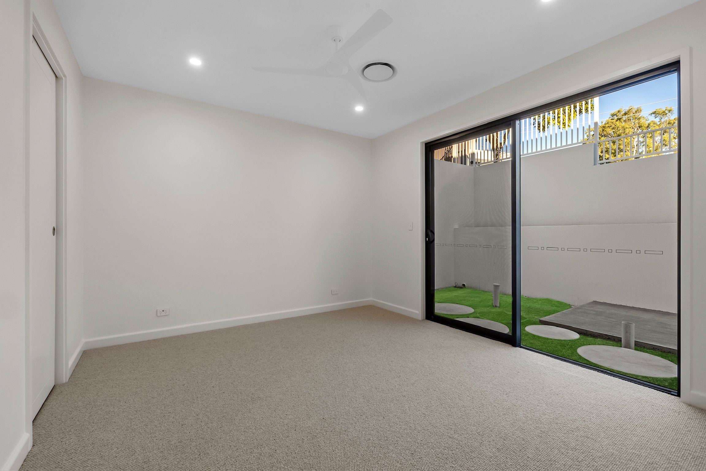 Empty room with beige carpet, white walls, ceiling fan, and black sliding glass door leading to small outdoor yard with artificial grass and stepping stones.