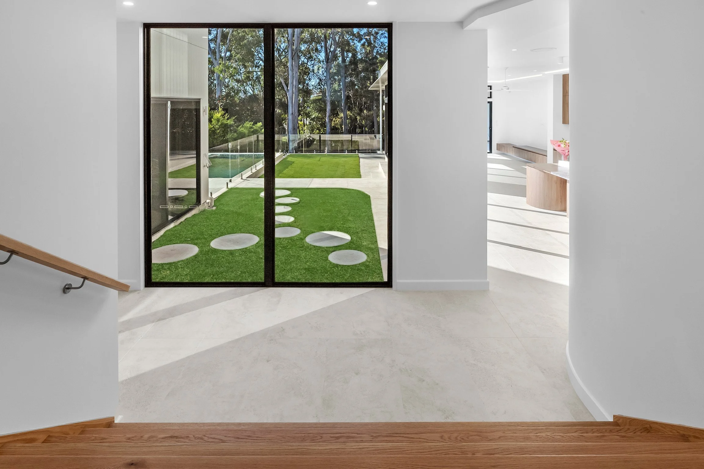 Interior view of a home with large sliding glass doors leading to a small backyard with artificial grass and trees outside. The space features white walls, light-colored flooring, and a wooden trim at the base of the walls. To the right, part of a ki