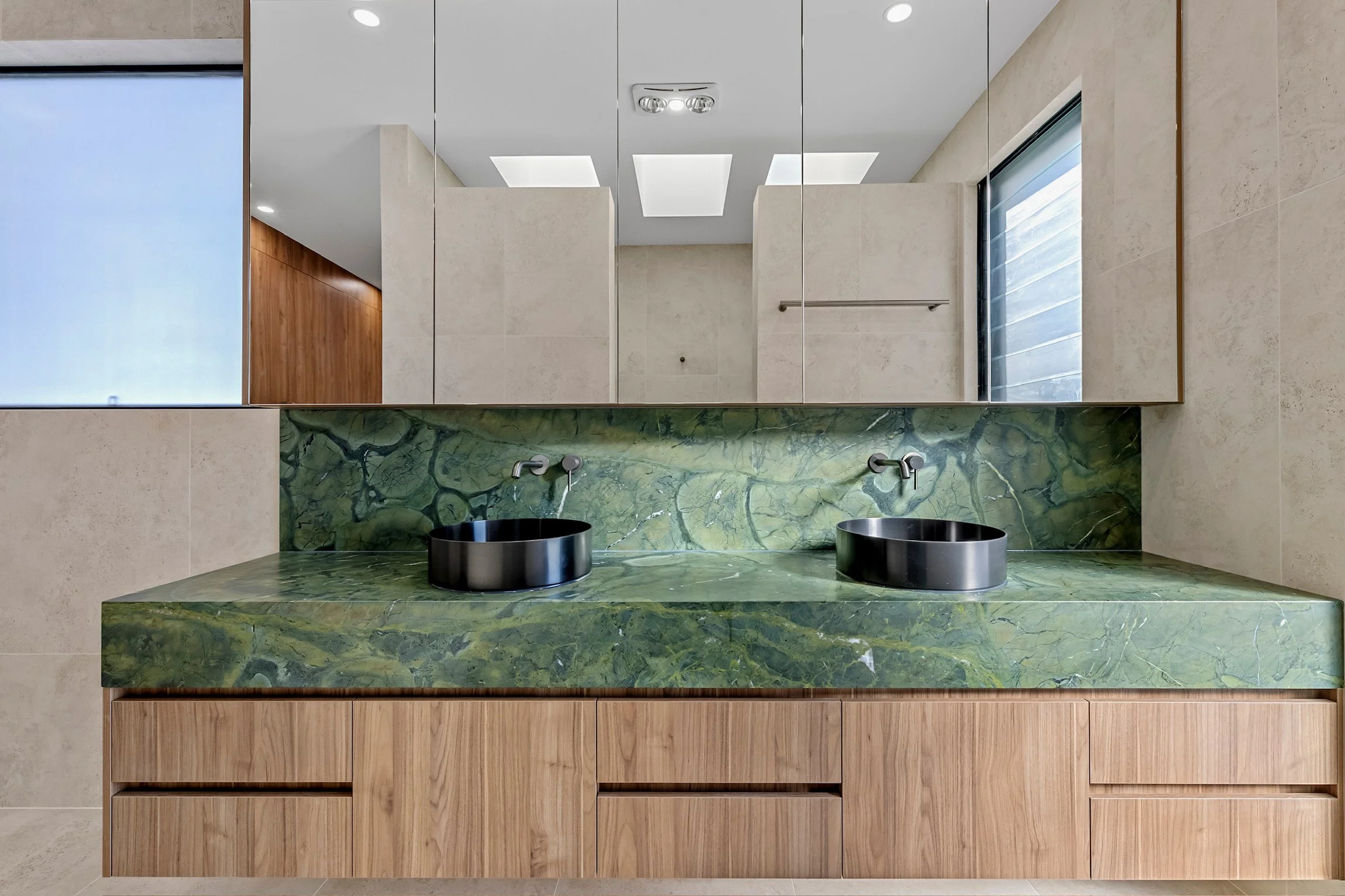 Modern bathroom vanity with a green marble countertop featuring two black vessel sinks and a large mirror above. The cabinet is made of light wood with open shelving and the walls are beige with a window on the right side.