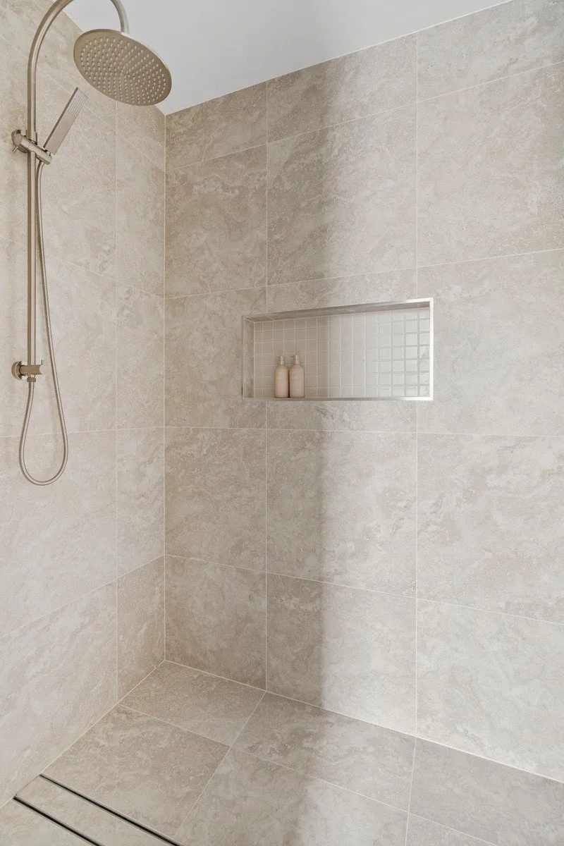 A beige tiled walk-in shower with a rain showerhead, a built-in wall niche with two bottles, and a linear floor drain.