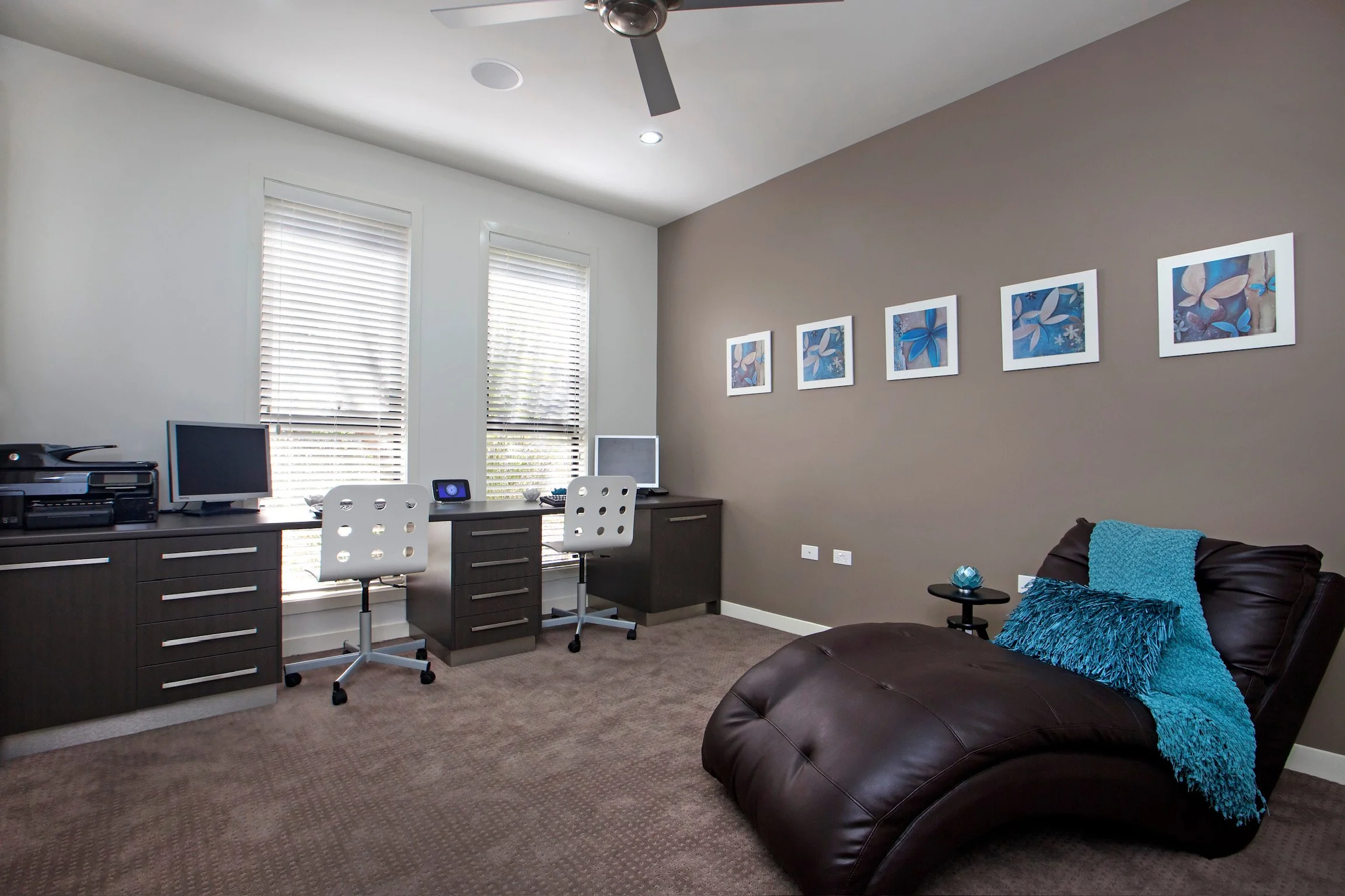 Home office with two desks, a computer, a printer, and framed artwork of blue flowers and butterflies on a beige wall. There are two windows with blinds, a brown leather armchair with a blue throw blanket, and a small side table with a decorative gla
