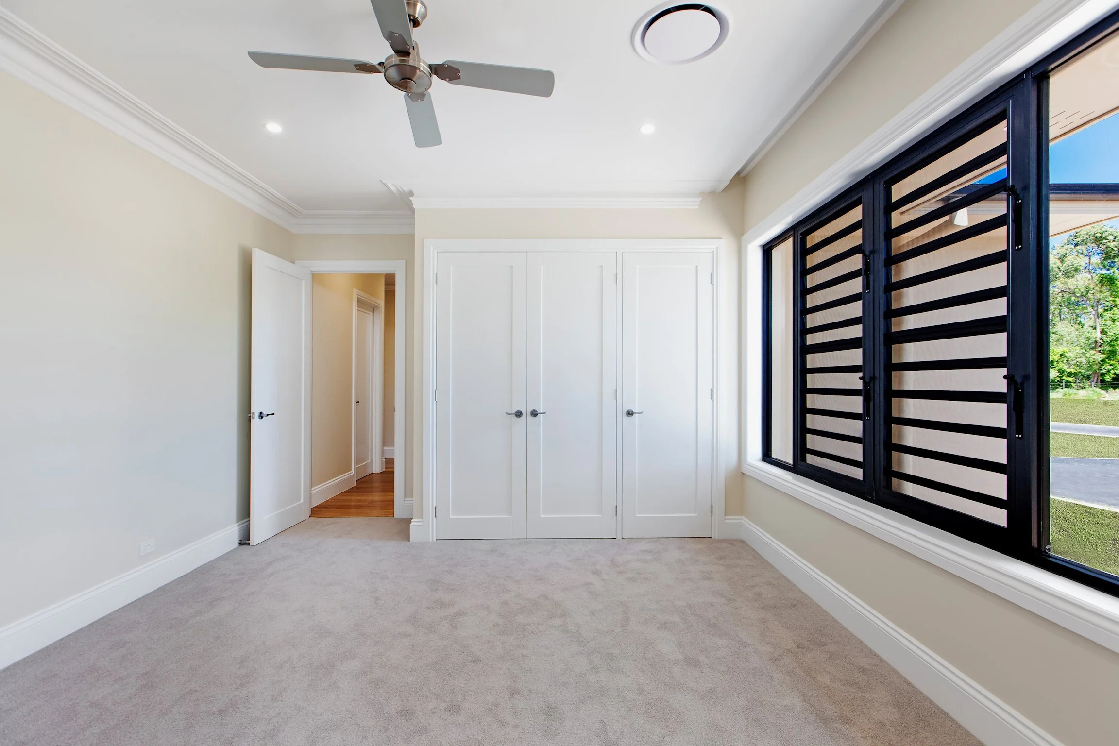 Empty room with beige walls, white trim, beige carpet, a ceiling fan, and windows with black security bars showing greenery outside.