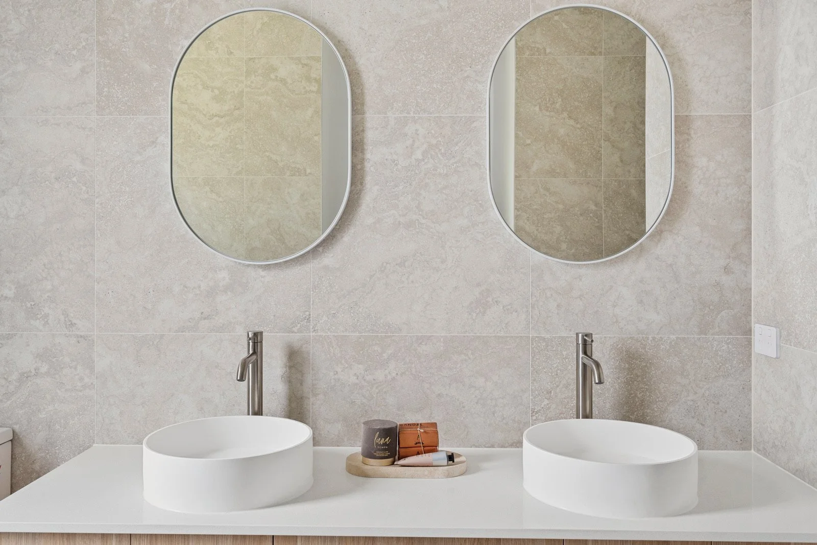 Modern bathroom double vanity with two oval mirrors, two vessel sinks, and a small tray with toiletries on the white countertop.