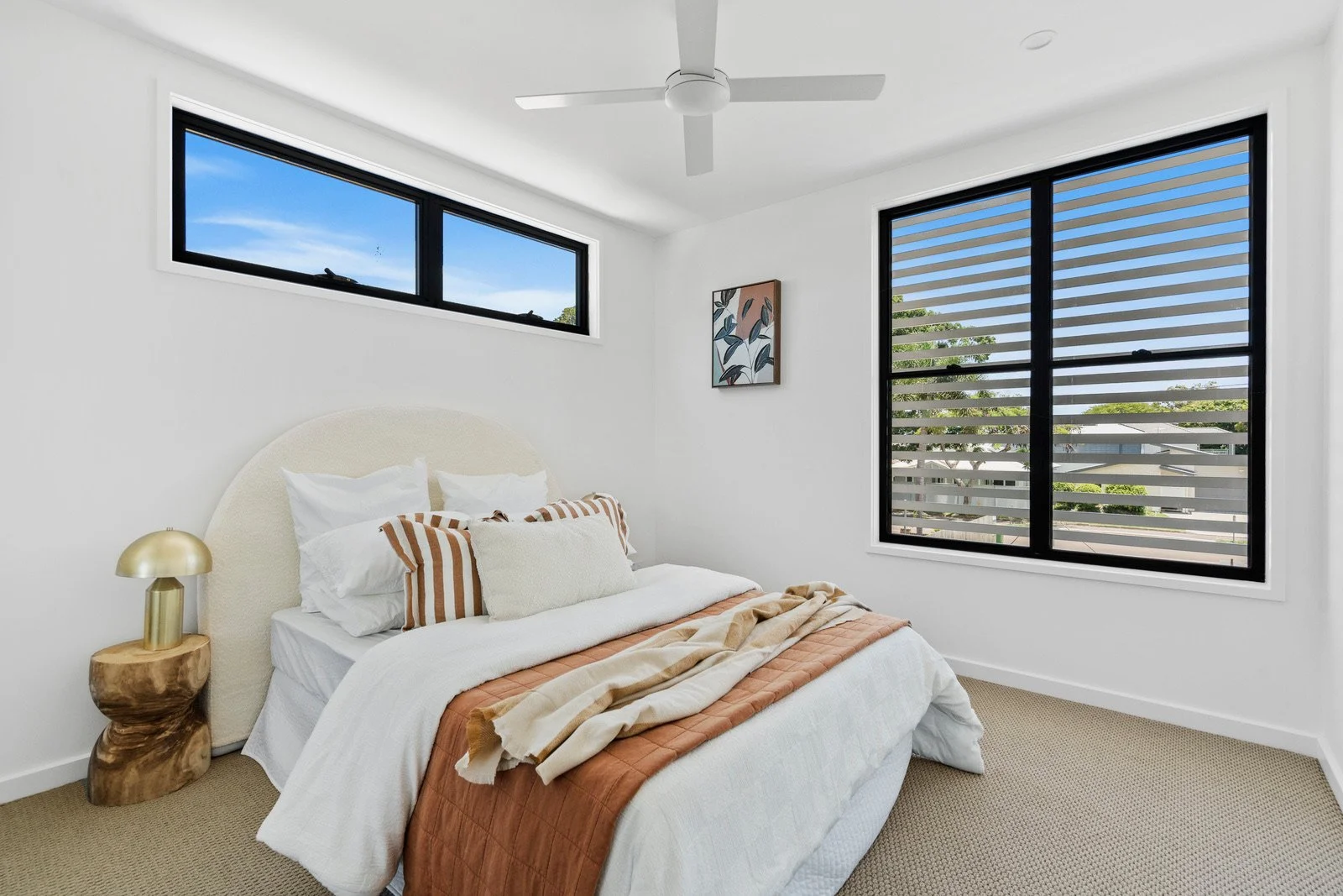 A bedroom with white walls, large windows with black frames, a white bed with white and patterned pillows, a beige headboard, a wooden nightstand with a gold lamp, and a piece of wall art. Outside the windows, a clear blue sky and greenery are visibl