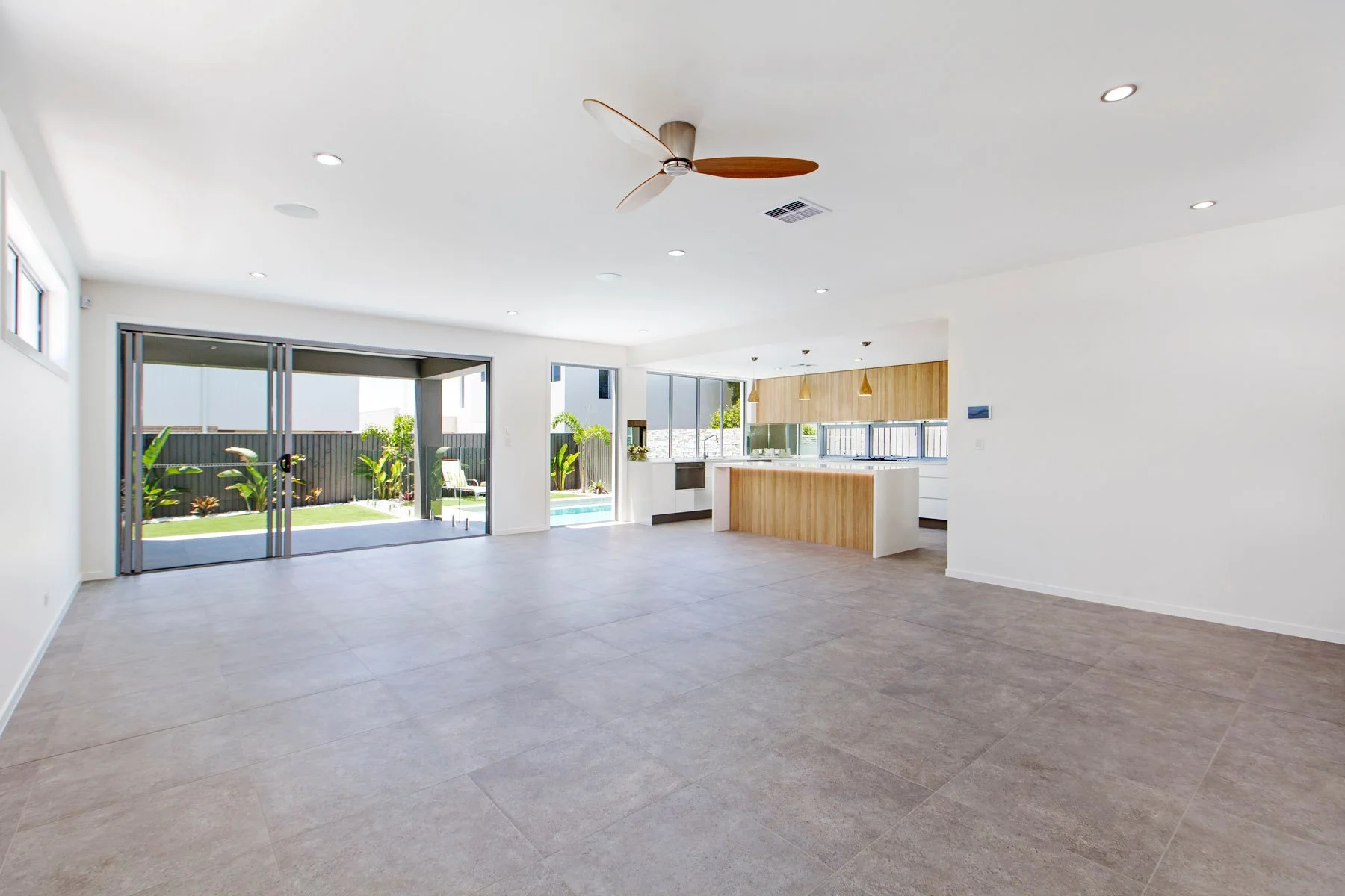 Bright, open living space with large sliding glass doors leading to a backyard with a small pool and plants. The room has a ceiling fan and recessed lighting, with a modern kitchen area featuring wooden accents.
