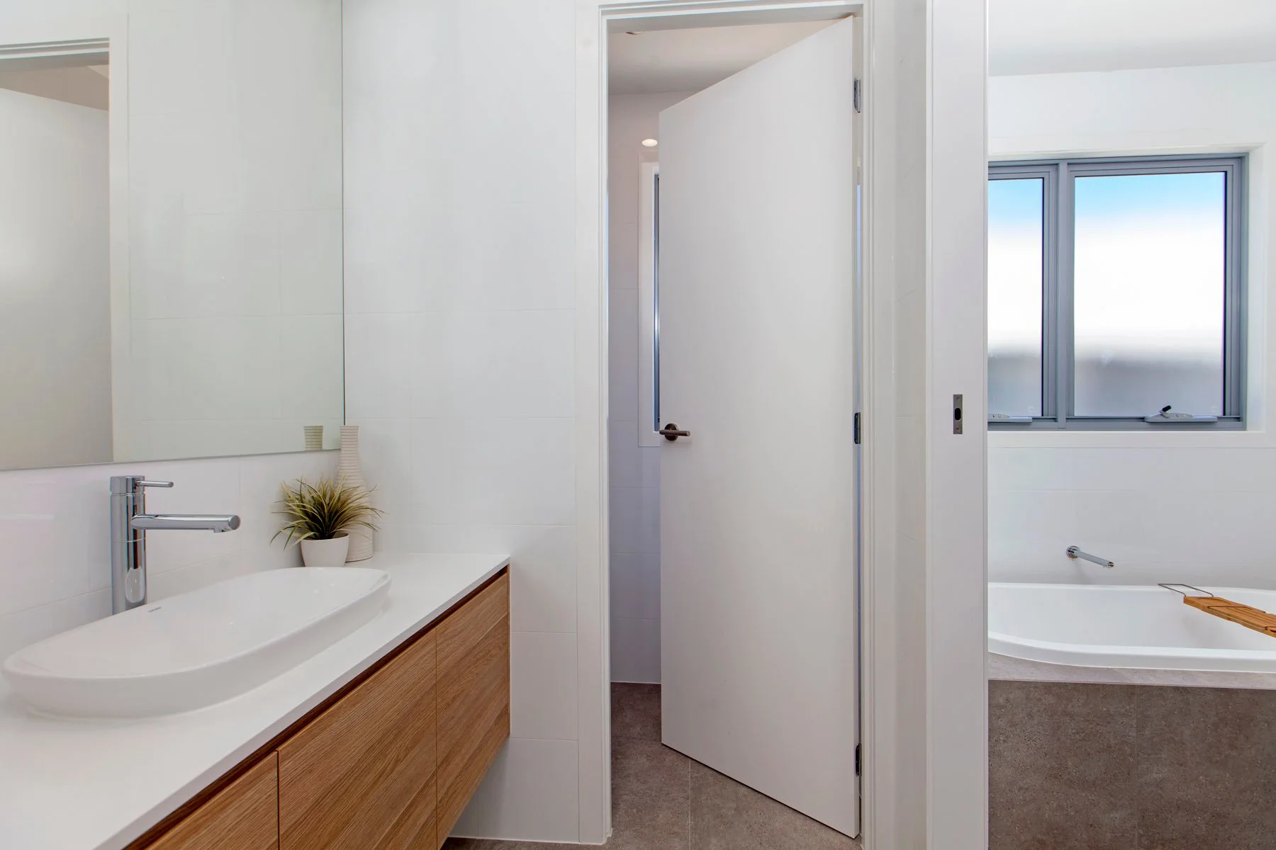 Modern bathroom with a white sink and wooden cabinet, a mirror, decorative vases with grass, a partially open door leading to a toilet room, a window with a view outside, and a bathtub with a wooden bath tray.