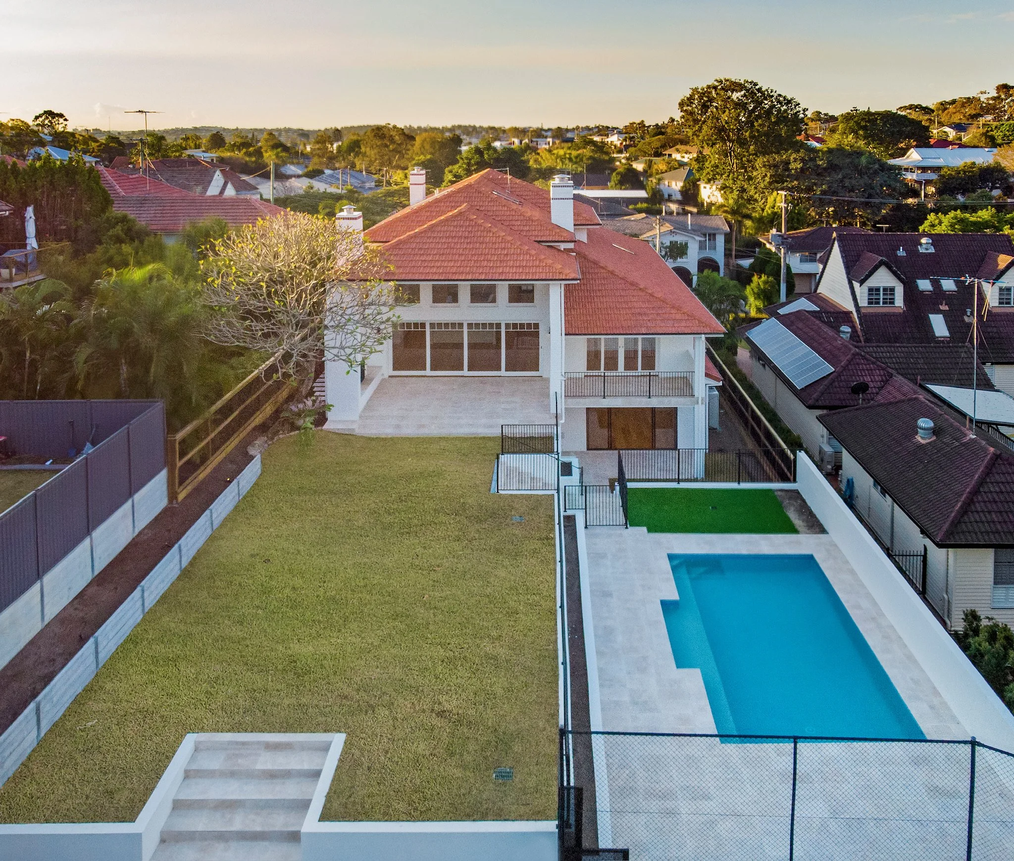 A backyard with a pool, a patio, a grassy lawn, and a modern house with large windows and a deck, surrounded by a black fence, with neighboring houses and trees in the background.