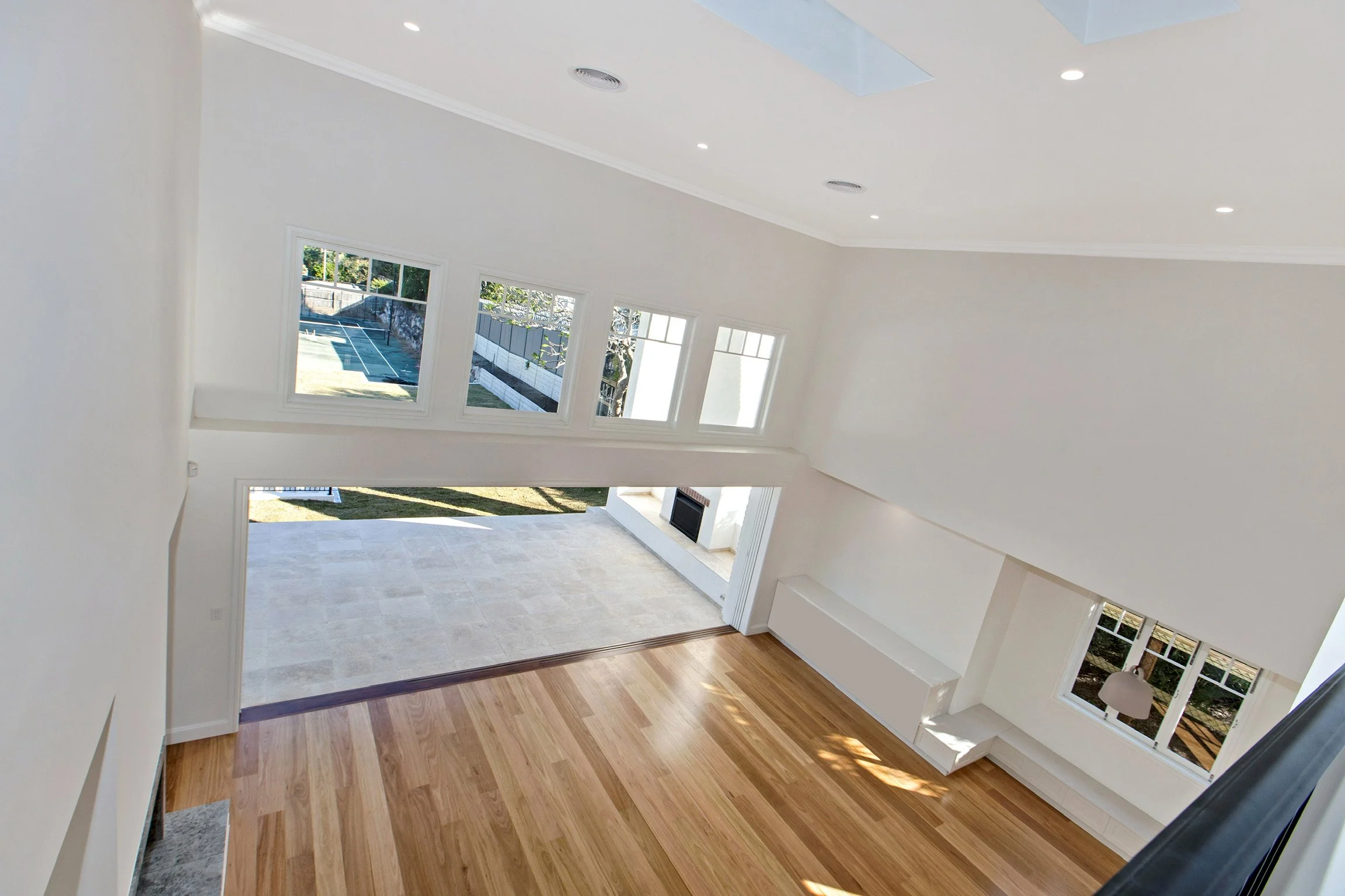 Empty modern living room with wooden flooring, large windows, and white walls, looking outside at a yard with a tennis court.
