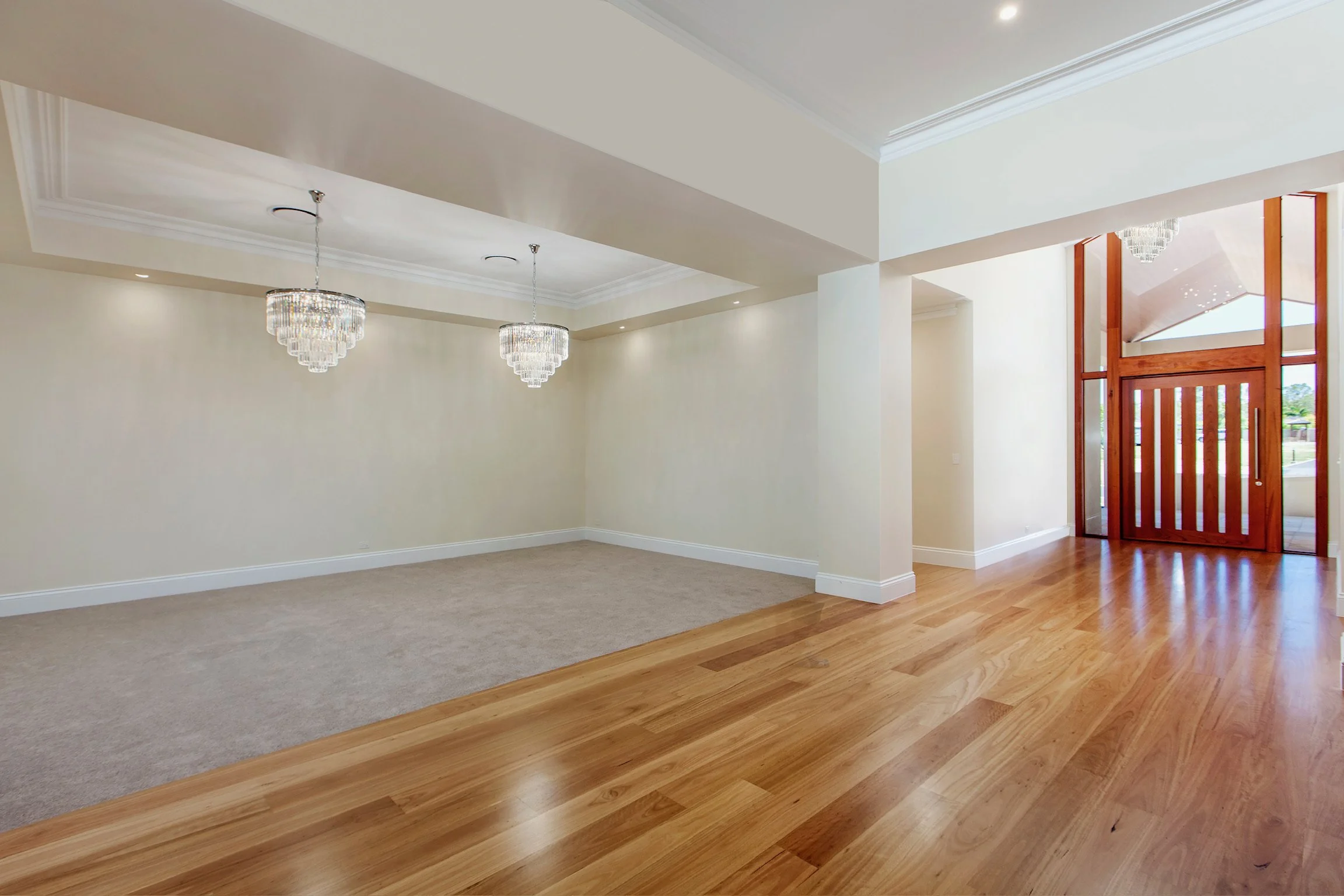 An empty living space with beige walls, white crown molding, and two crystal chandeliers hanging from the ceiling. The room has a carpeted area on the left and hardwood flooring on the right, with a wooden and glass front door on the right side.