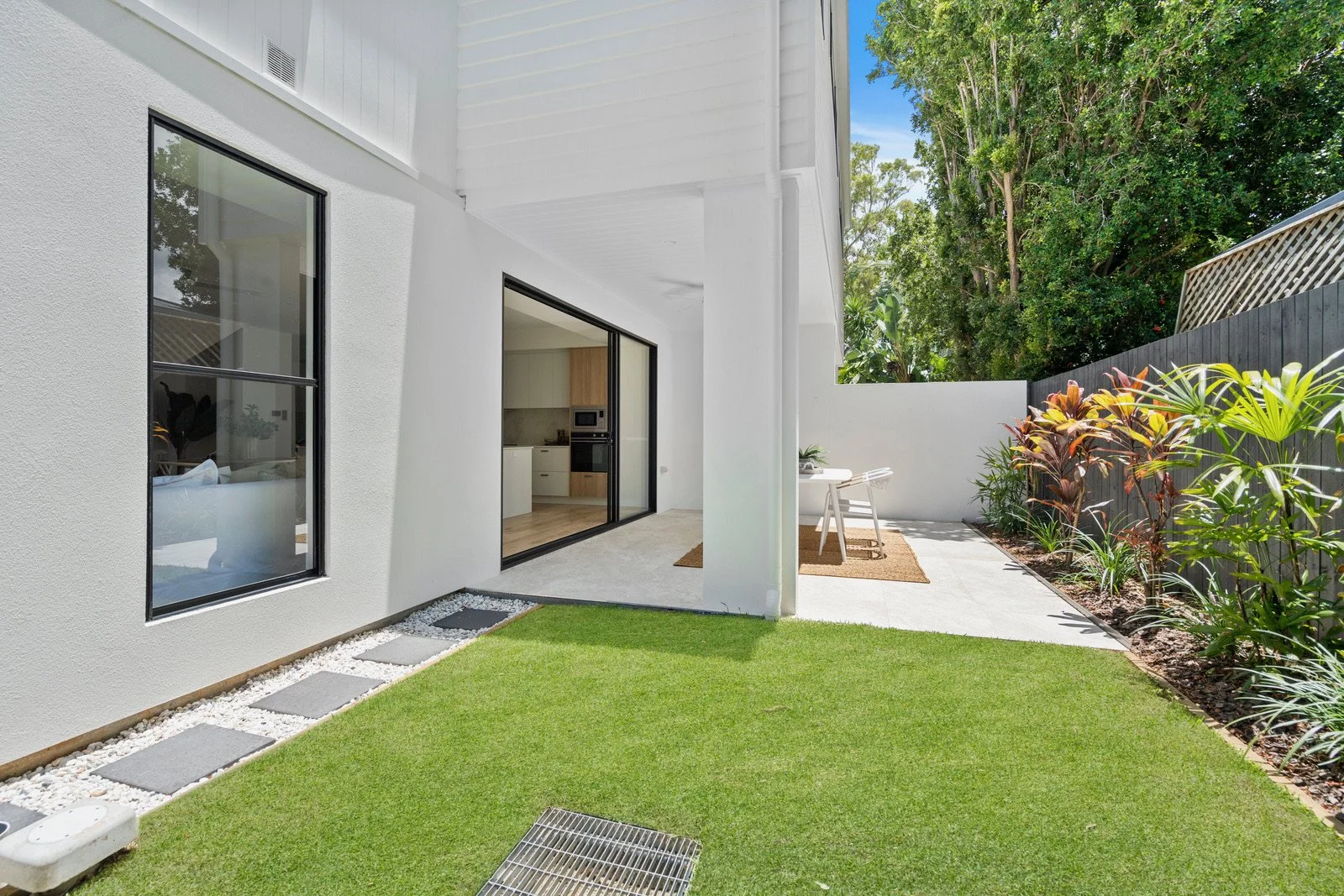 Modern backyard with green lawn, white exterior house wall with large black-framed windows, sliding glass door open to interior kitchen, small patio with outdoor table and chairs, landscaped garden with plants along a black fence, and tall trees in t