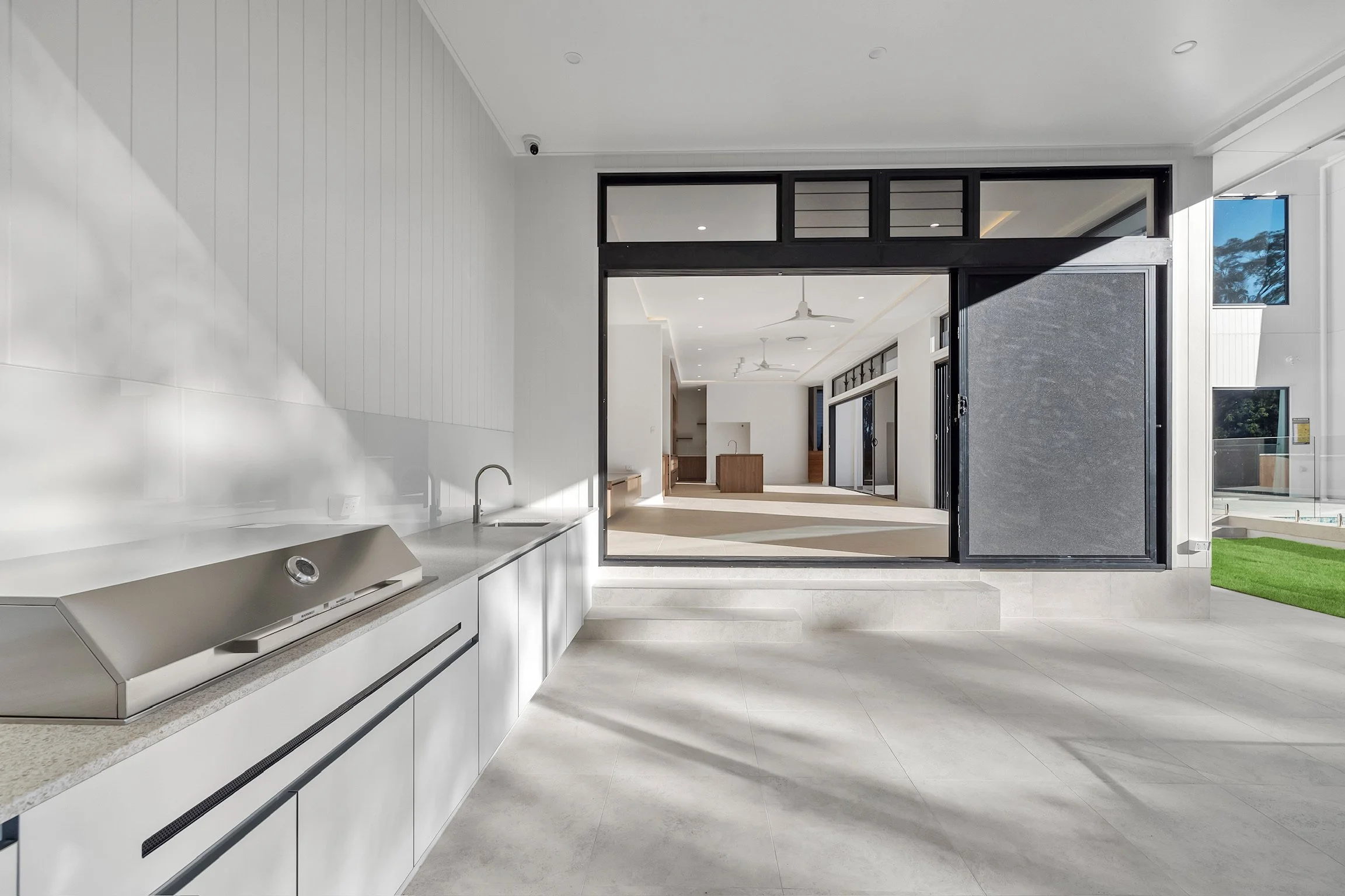 Modern kitchen with white cabinets, built-in oven, light-colored tile flooring, large sliding glass doors opening to a patio, and a view of the interior living space with ceiling fans.