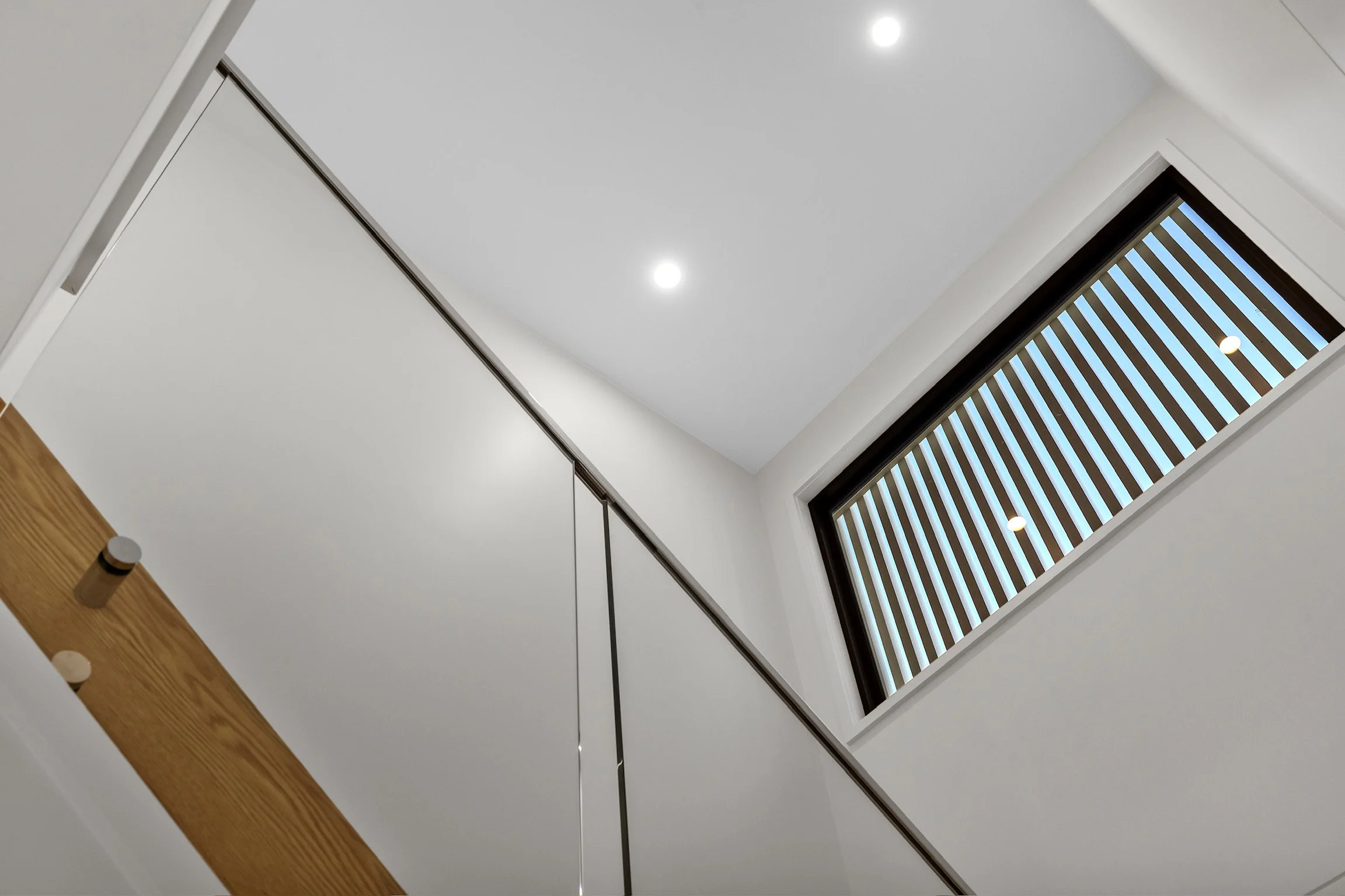An interior view looking up at a ceiling with two bright recessed lights, a window with wooden blinds, and a section of wood paneling on the wall.
