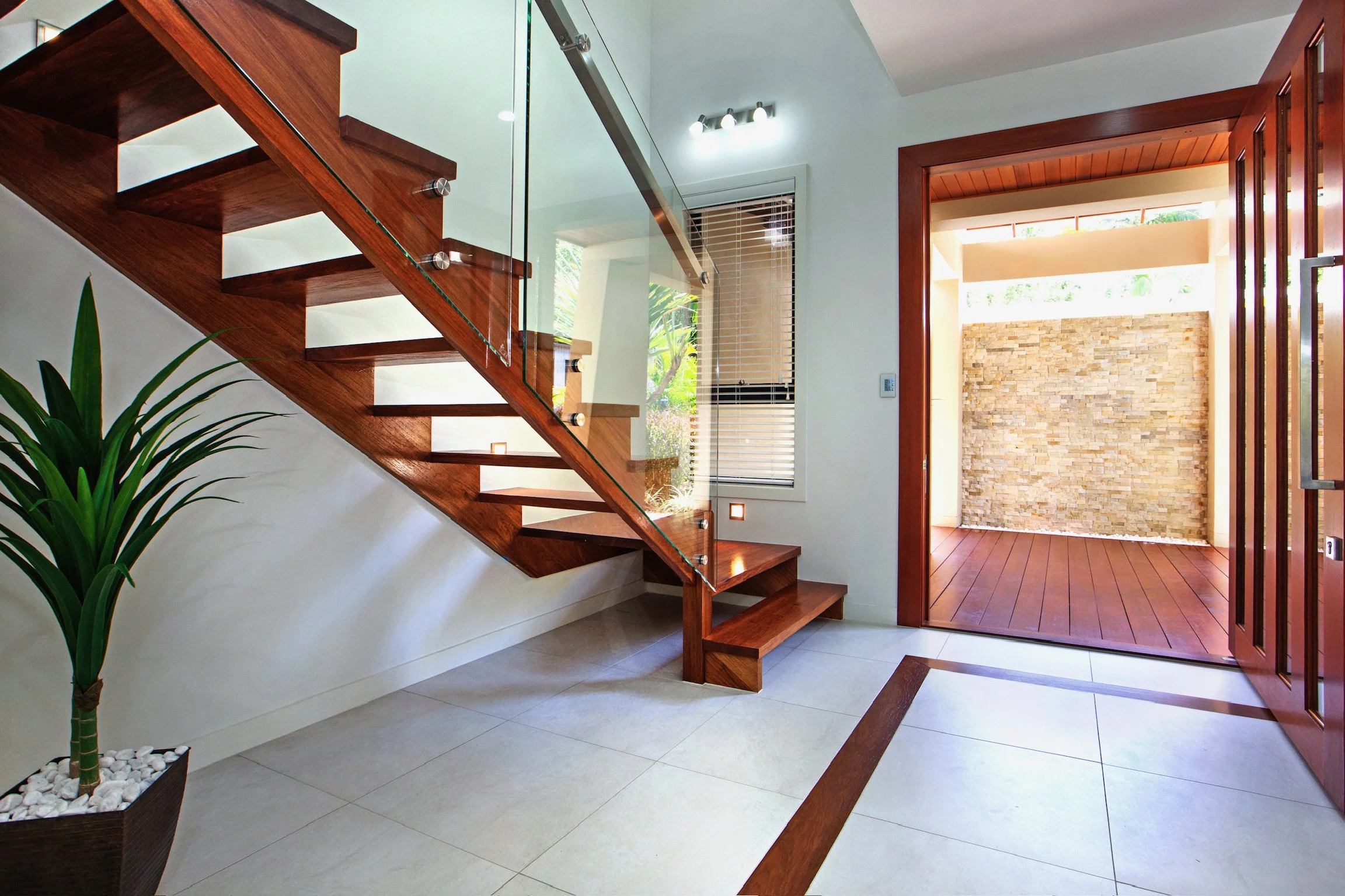 Modern interior entrance area with wooden staircase featuring glass railing, potted plant with white stones, large window with blinds, and open wooden door leading to a room with stone accent wall.