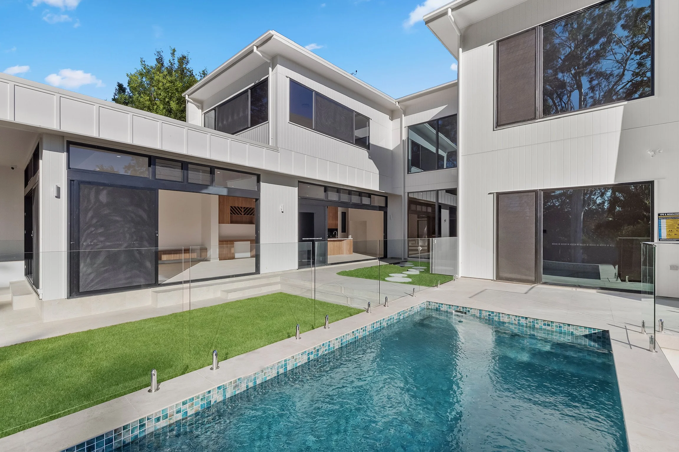 Modern white multi-story house with large windows and a backyard pool surrounded by a glass fence, green lawn, and stepping stones.