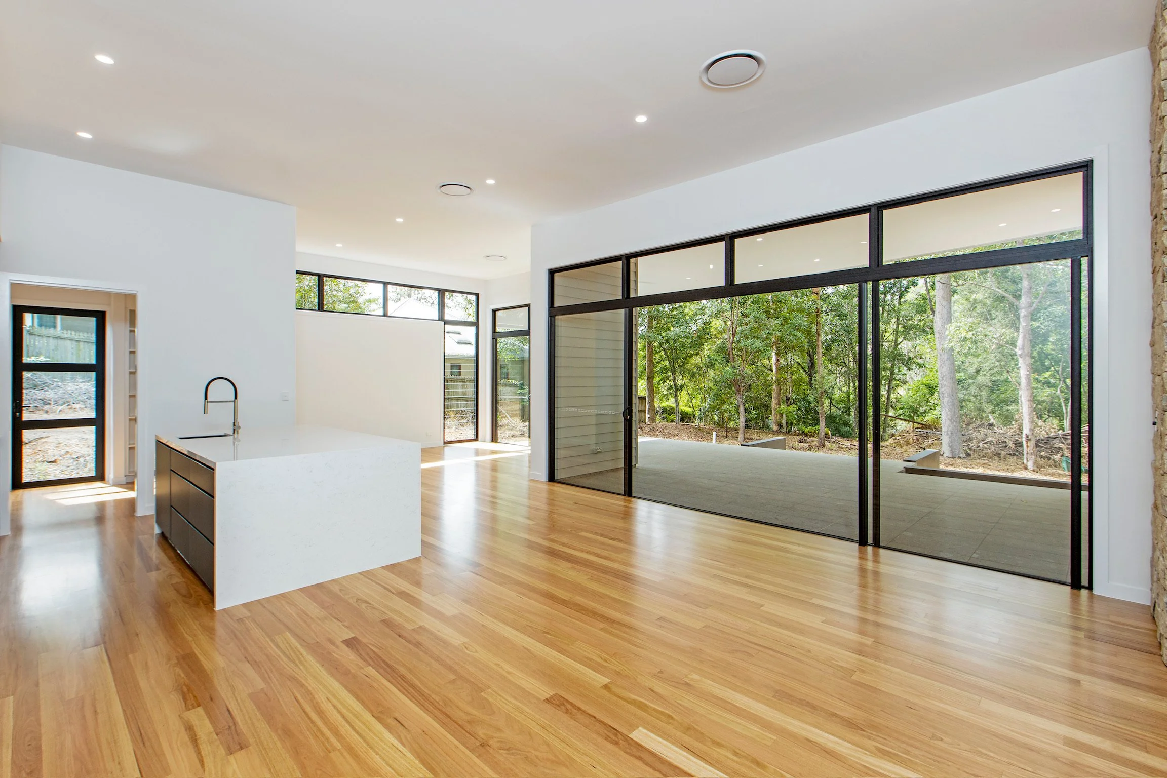 Open-concept living area with hardwood floors, white walls, large sliding glass doors showing a wooded outdoor view, and ceiling lights.