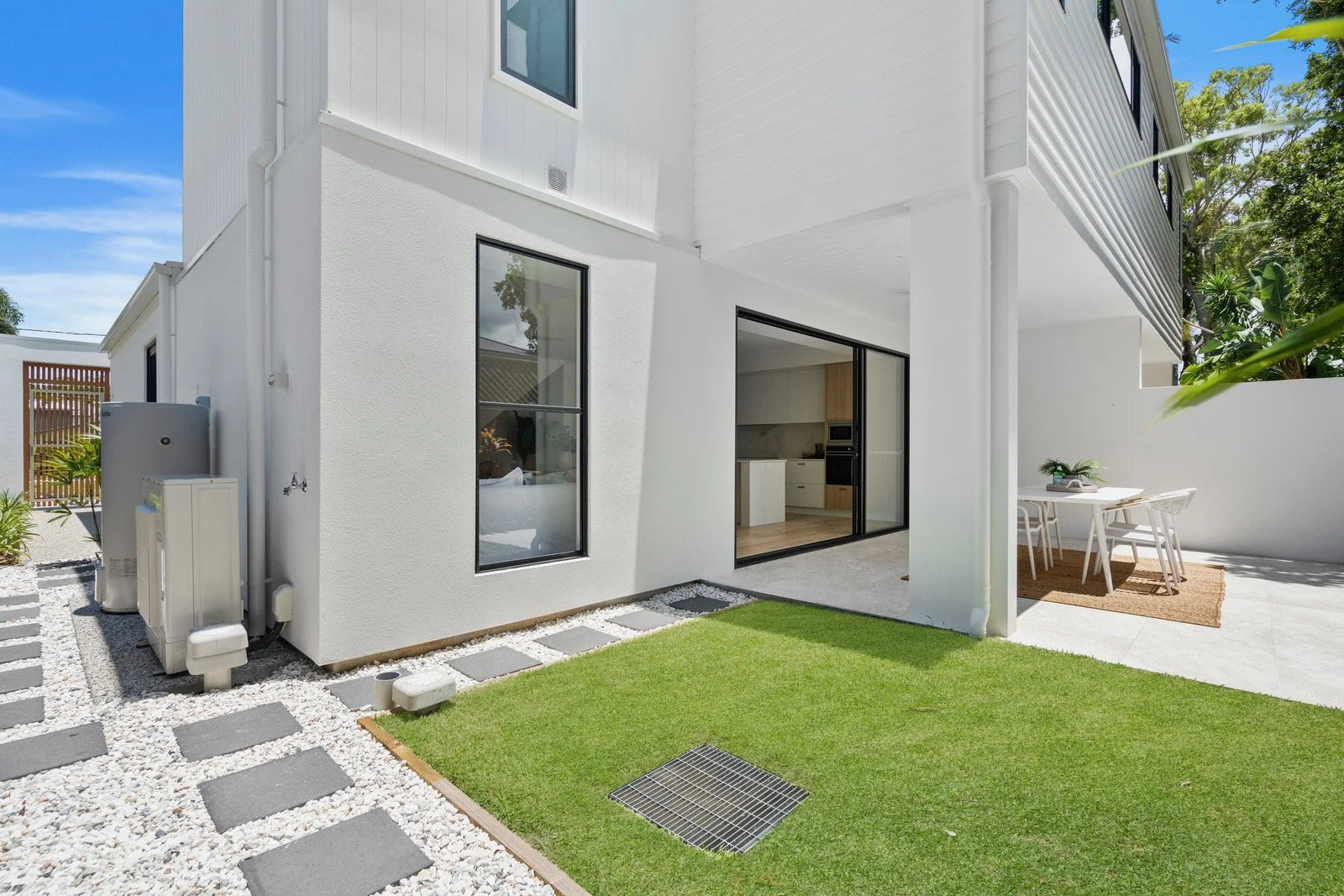 Modern white house with large sliding glass doors, a small backyard with green grass, outdoor dining area, and trees in the background.