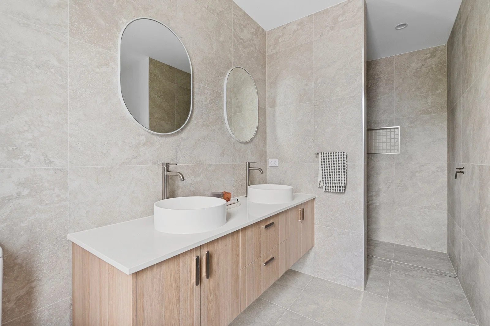 Modern bathroom with dual vessel sinks, oval mirrors, beige tiled walls and floor, wooden vanity, towel rack with checkered towel, and walk-in shower with tiled walls and a recessed shelf.